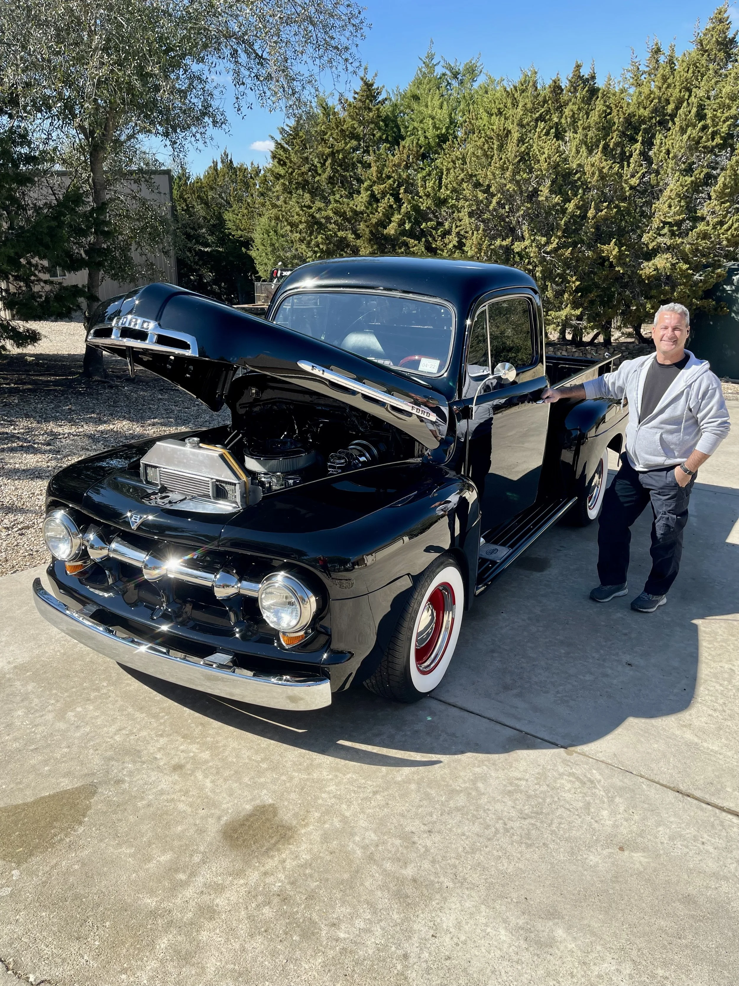 A man standing next to a vintage black Ford pickup truck with the hood open, revealing the engine, parked on a concrete driveway with trees in the background.