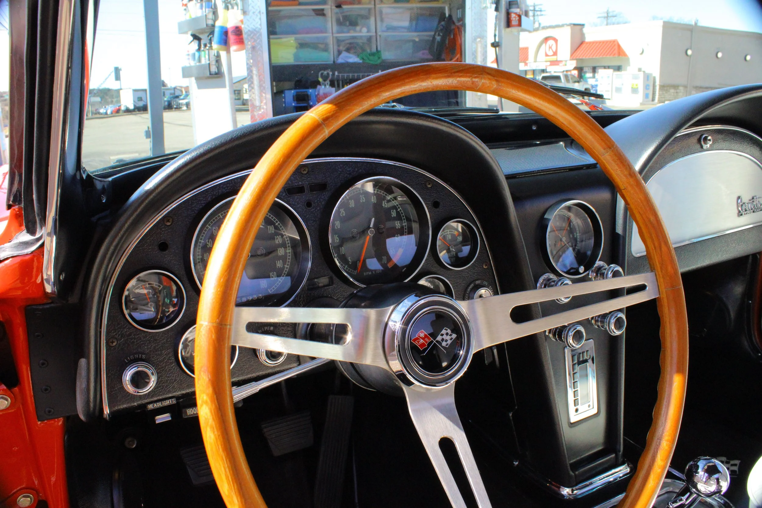 Close-up of a classic car dashboard and steering wheel, showing speedometer, tachometer, and other gauges, with a wood-rimmed steering wheel and a black dashboard.