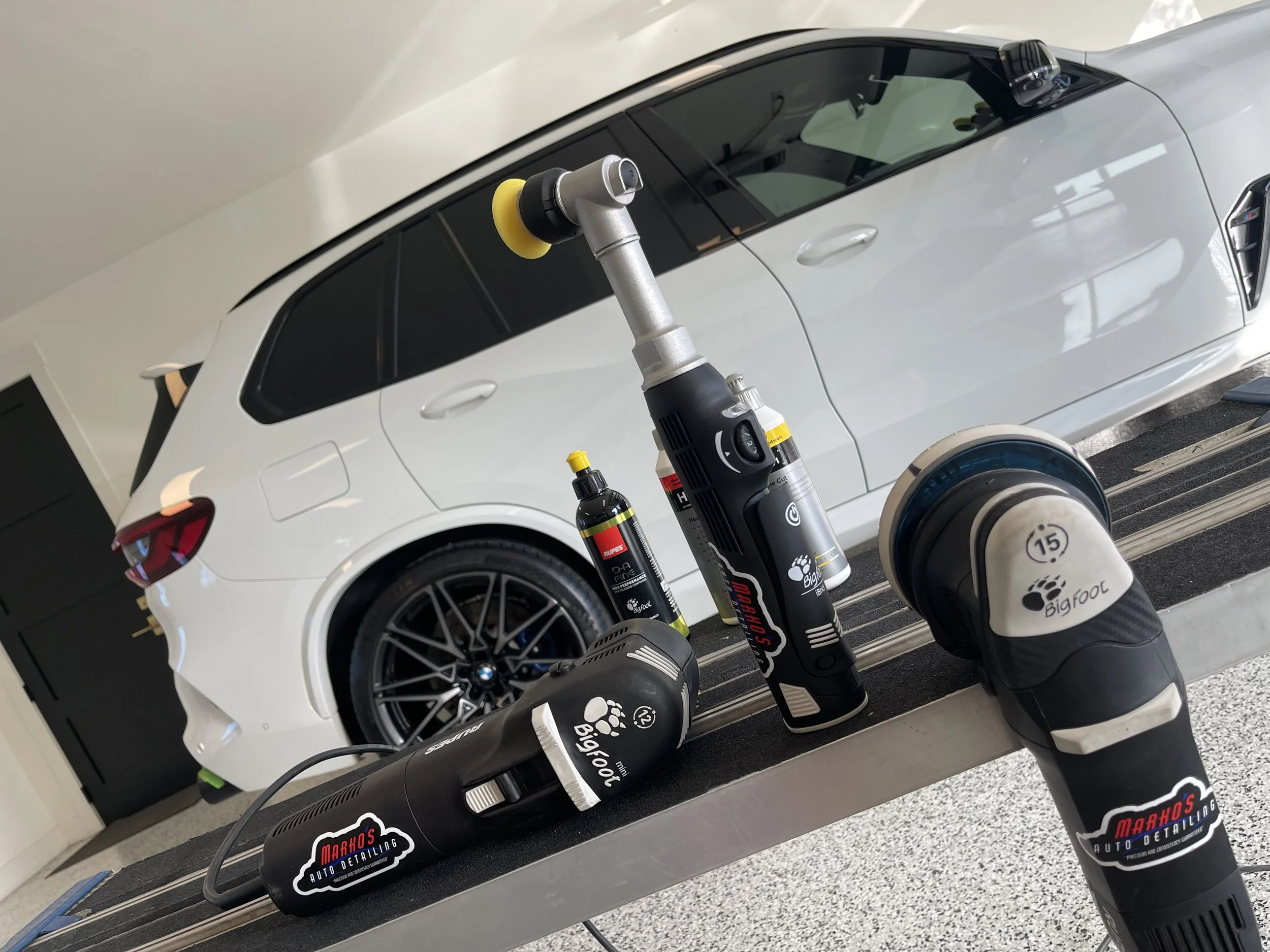 A white BMW car in a garage or detailing shop with various car detailing tools and polishers on a black work surface in the foreground.