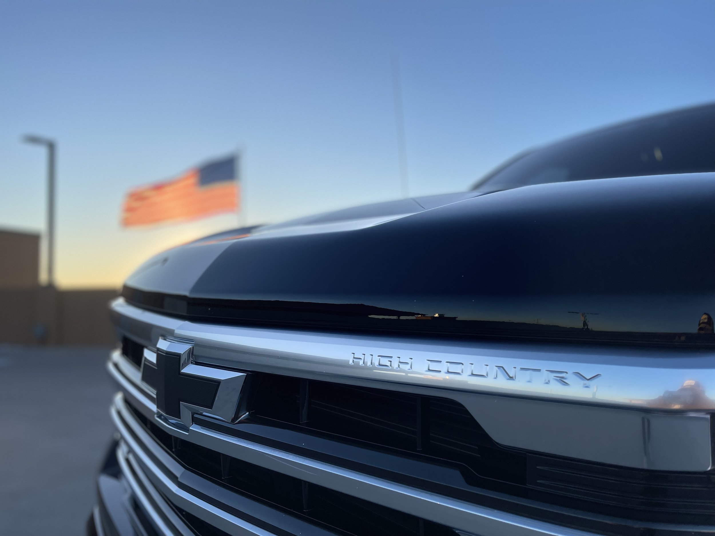 Close-up of the front grille of a Chevrolet vehicle with a Chevrolet emblem and the words 'High Country,' with an American flag in the background at sunset.