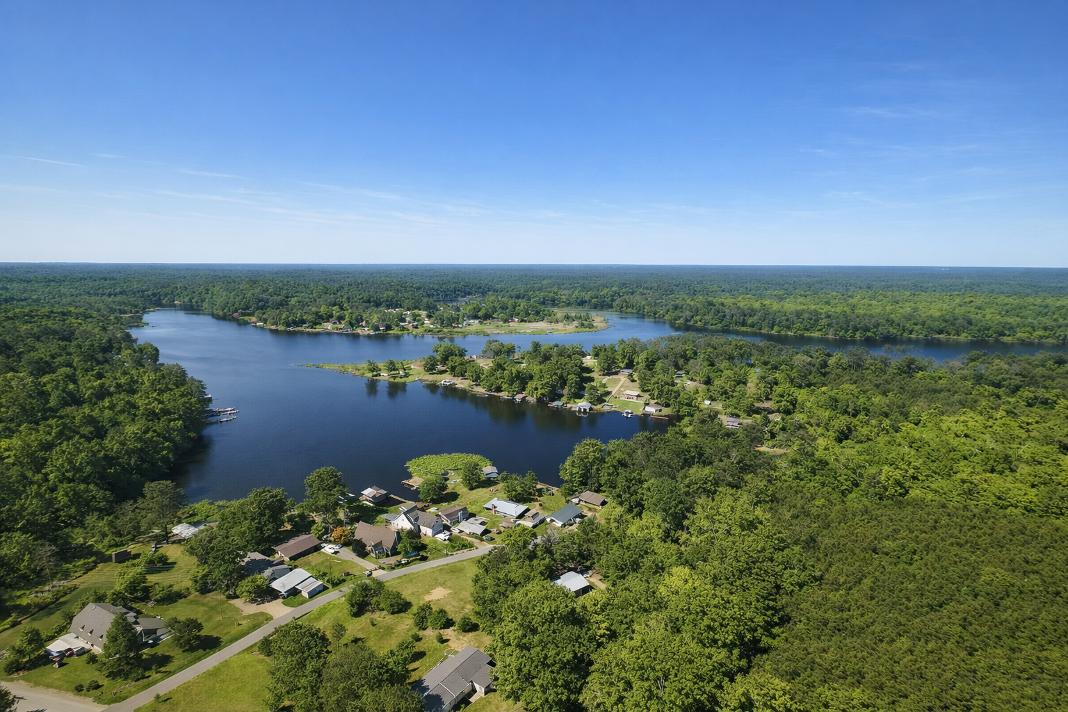 Aerial view of a lake surrounded by a mix of houses, docks, and dense green forest under a clear blue sky.