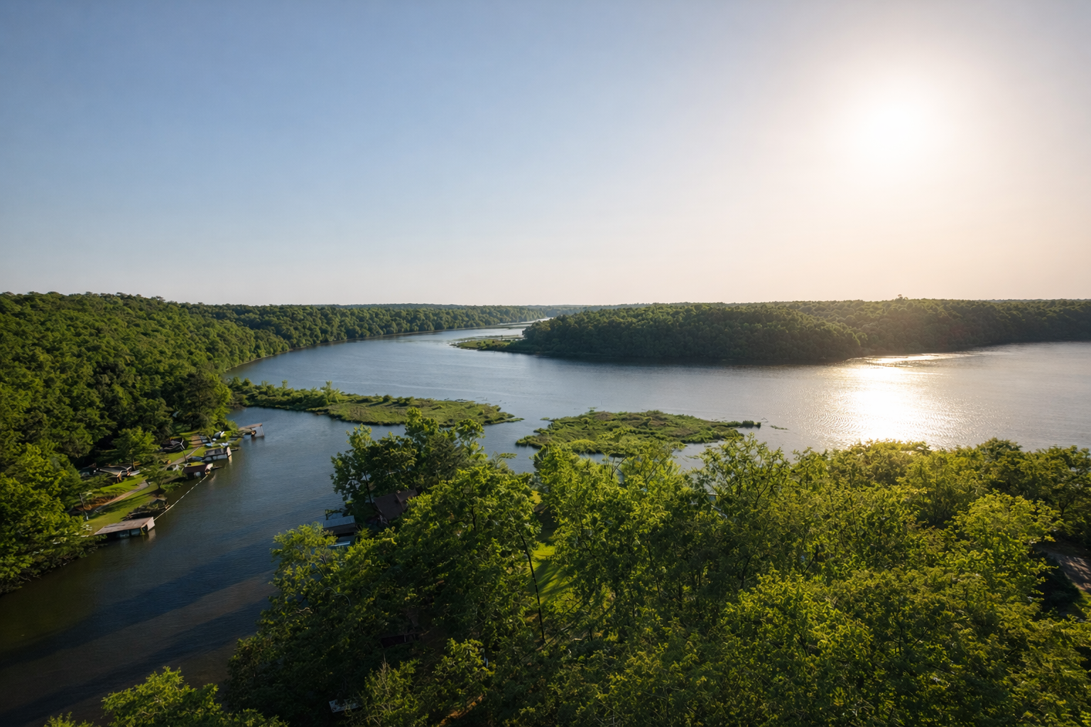 A panoramic view of a winding river flowing through a lush green landscape during sunset, with a few houses along the shore and dense trees covering the area.