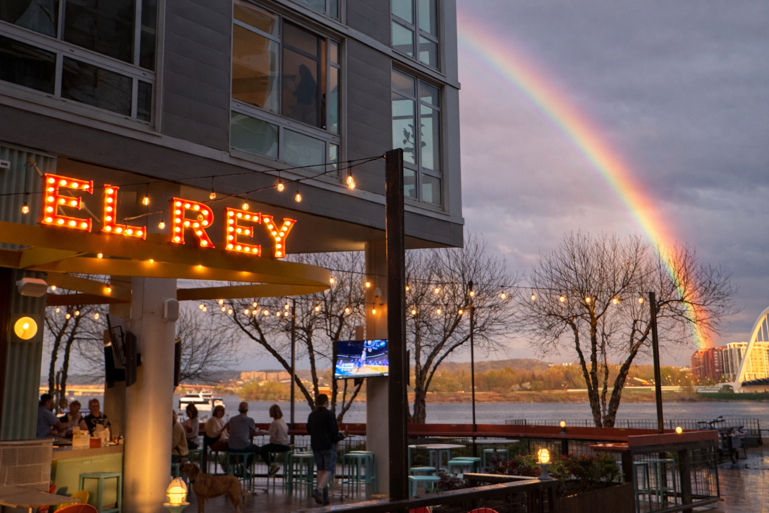 An outdoor restaurant and bar called El Rey with customers seated at tables. There are string lights, a rainbow in the sky, and some leafless trees near a body of water. Located in Navy Yard SE near the Washington Nationals baseball park.