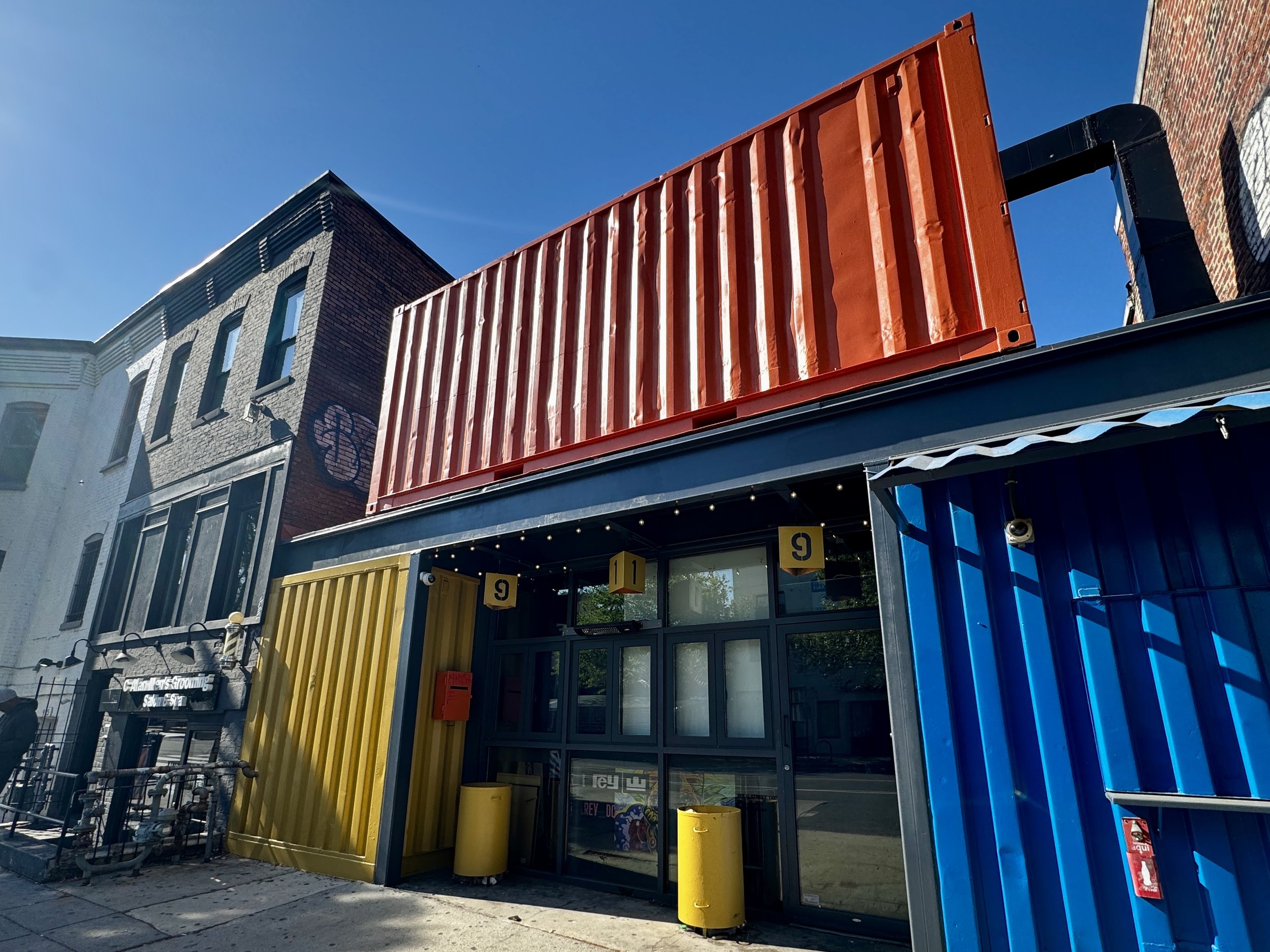 Colorful bar storefront made of shipping containers with red, yellow, and blue sections, adjacent to a brick building with arched windows, under a clear blue sky. On U Street NW Washington DC.