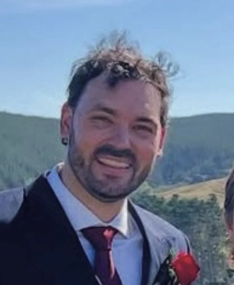 Richard Belcher from The Albion Club. A smiling man with light brown hair and a beard, wearing a suit, white shirt, and a red tie, standing outdoors against a hilly landscape with a blue sky.