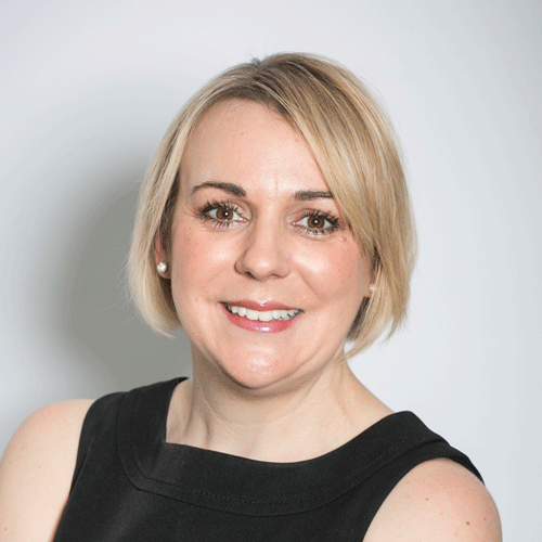 Kate Miller from Chilli Fox events. Portrait of a smiling woman with short blonde hair and pearl earrings, wearing a sleeveless black top against a light gray background.