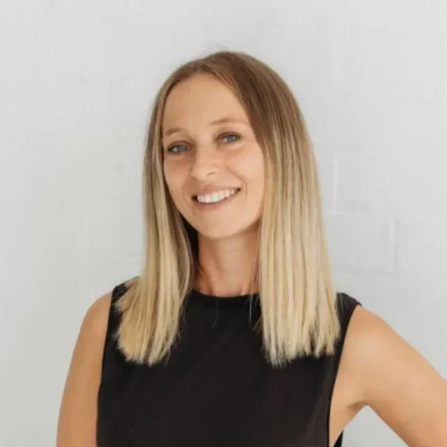 Eleanora Castellini from Tiny Yogis. A woman with blonde hair wearing a black sleeveless top, smiling, standing against a white wall.