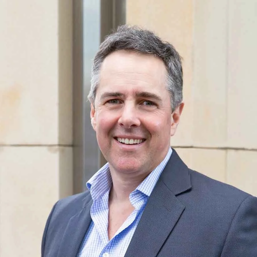 Ben Cowan from CCA Partners. Professional man with gray hair smiling, wearing a dark suit and blue dress shirt, standing outside against a beige building.