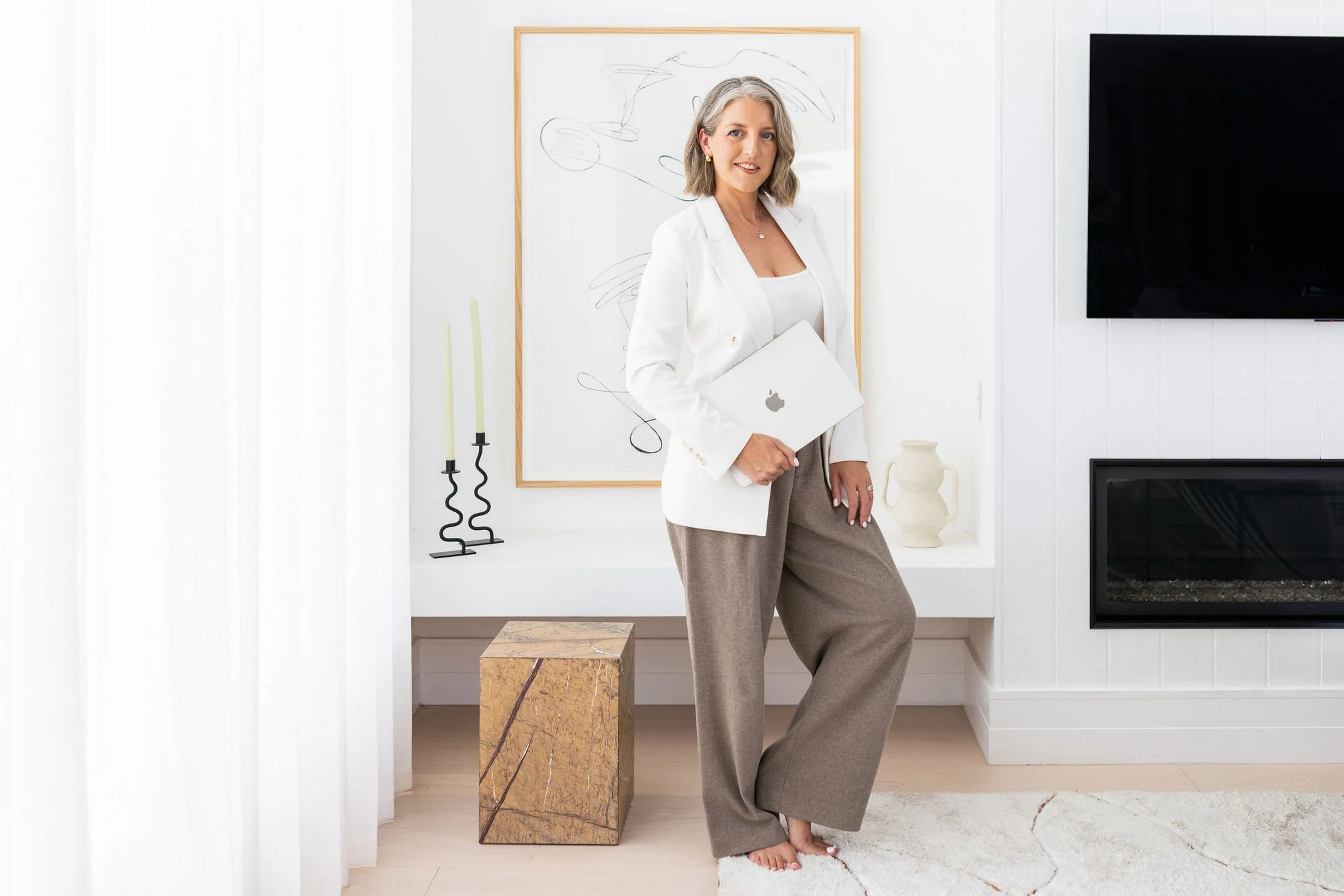 Katie Lodge from The Creative Lodge is pictured. She is a woman in a white blazer and beige pants standing in a bright, modern living room holding a closed MacBook laptop.