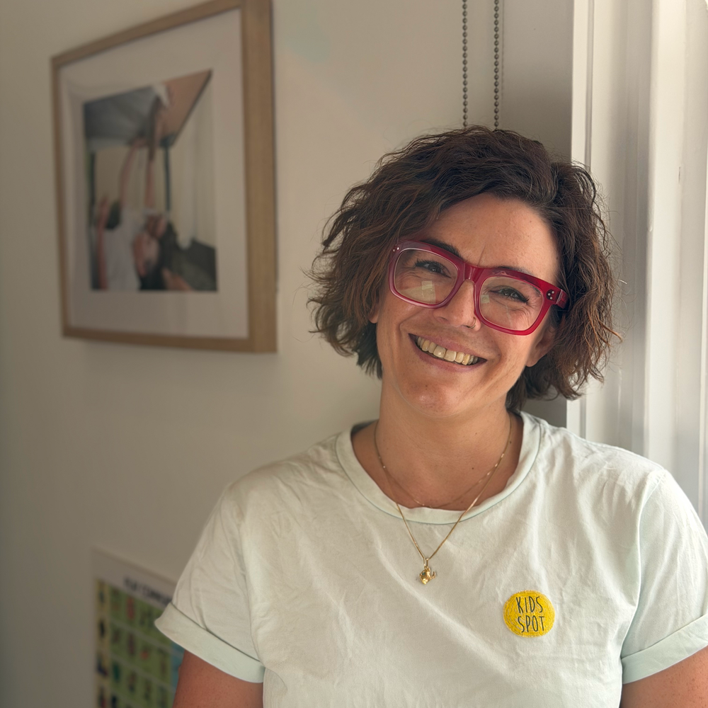 A woman with short, curly brown hair wearing red glasses and a white t-shirt with a yellow "Kids Spot" patch, smiling and standing indoors near a window with framed pictures on the wall.