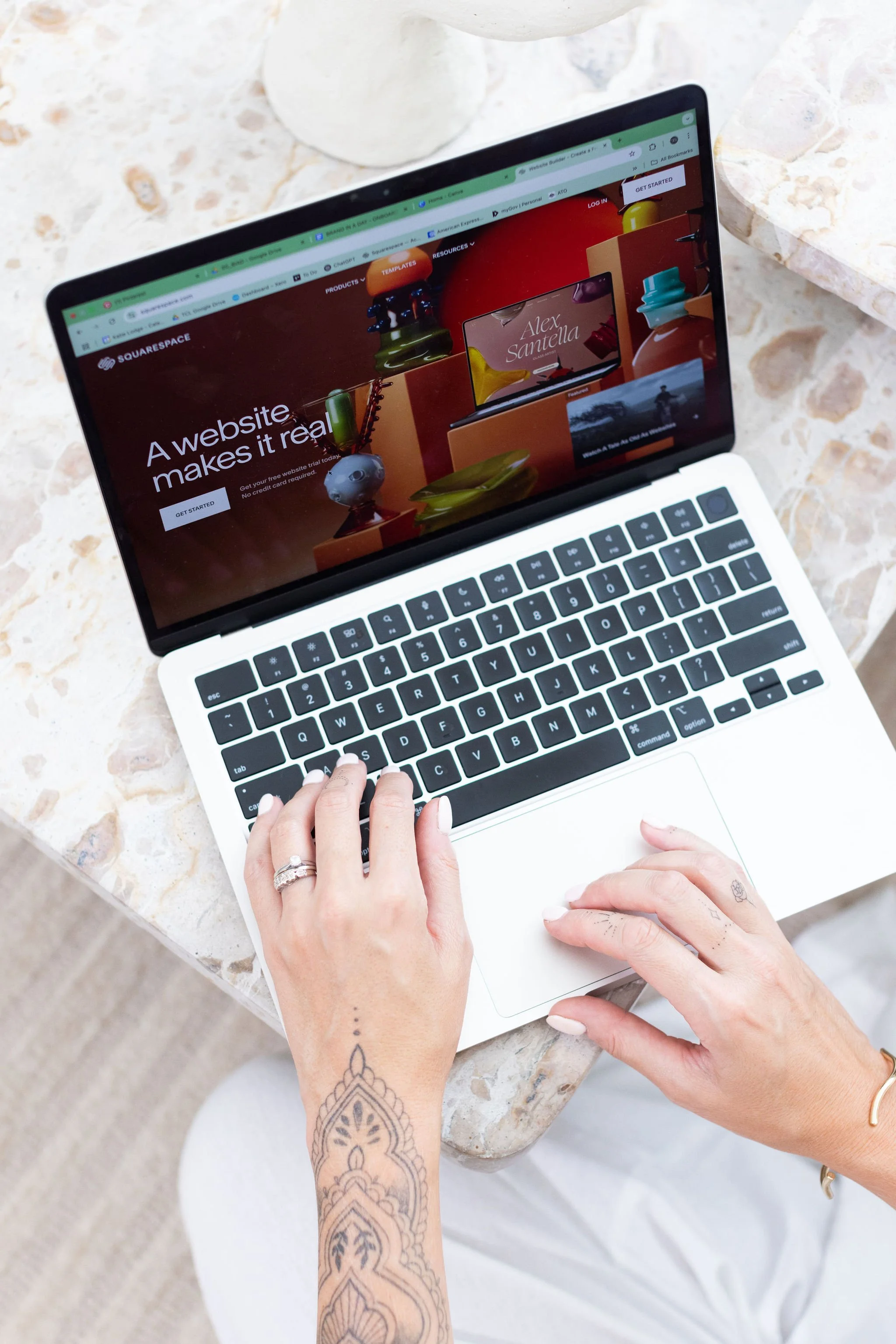 A laptop on a stone table displaying the Squarespace website with the message "A website makes it real." A person's hands are on the keyboard and trackpad, with tattoos on their arm and rings on their fingers.