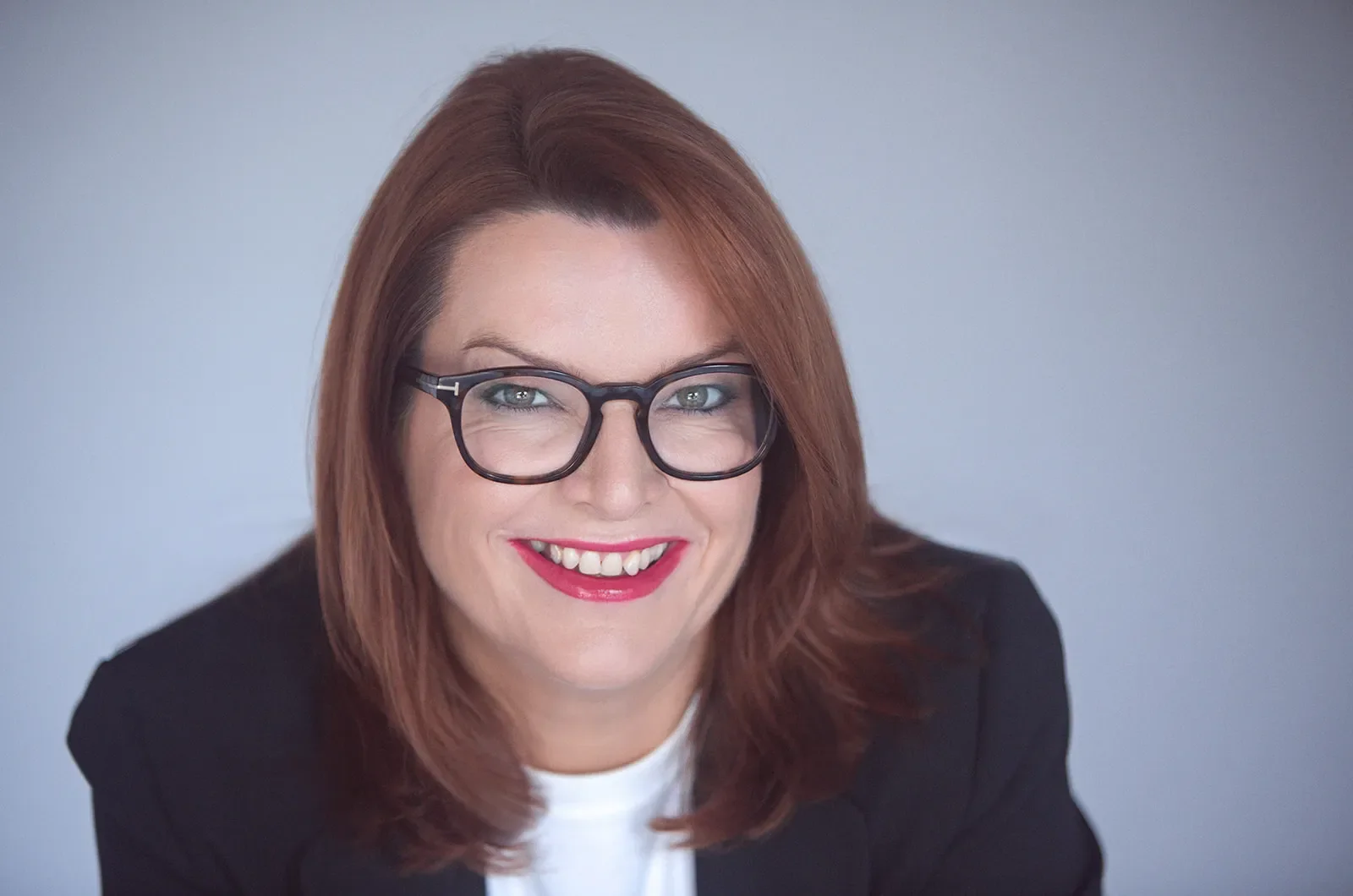 Portrait of a smiling woman with red hair and black glasses, wearing a black blazer and white shirt, against a plain light background.