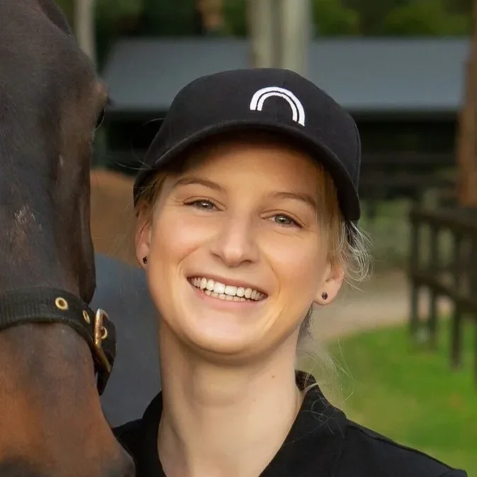 Meghan Andrew from Growth Edges. A young woman smiling, wearing a black cap and black shirt, standing next to a horse outdoors.