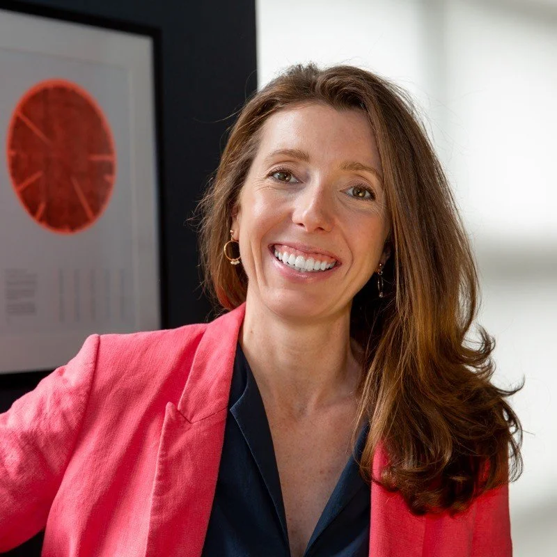 Pippa Corry from Philo&Co. A woman with long reddish-brown hair smiling and wearing a red blazer over a navy blue top, standing in an office setting with a framed chart or diagram in the background.