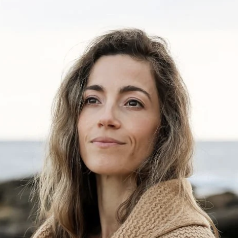 A woman with wavy brown hair standing outdoors near the water, wearing a tan sweater.