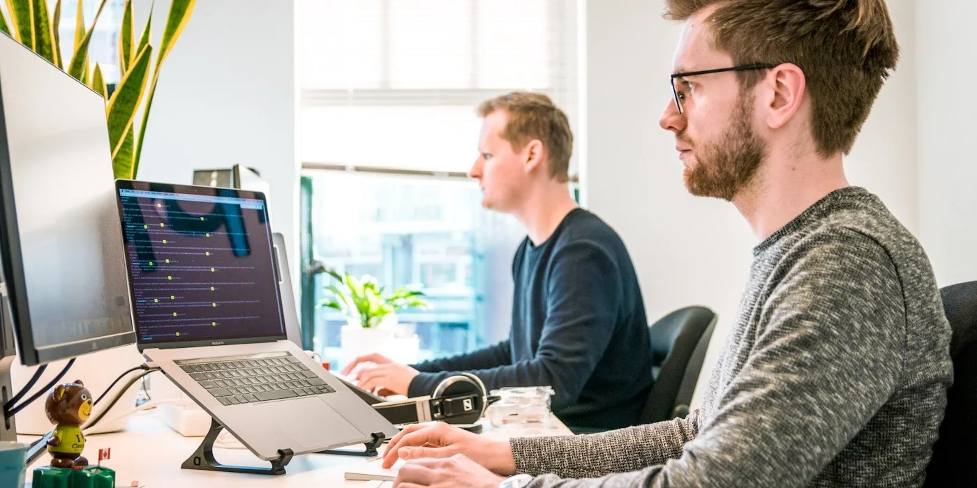 Two men working at their desks in an office, focusing on their computers, with a plant and windows in the background.