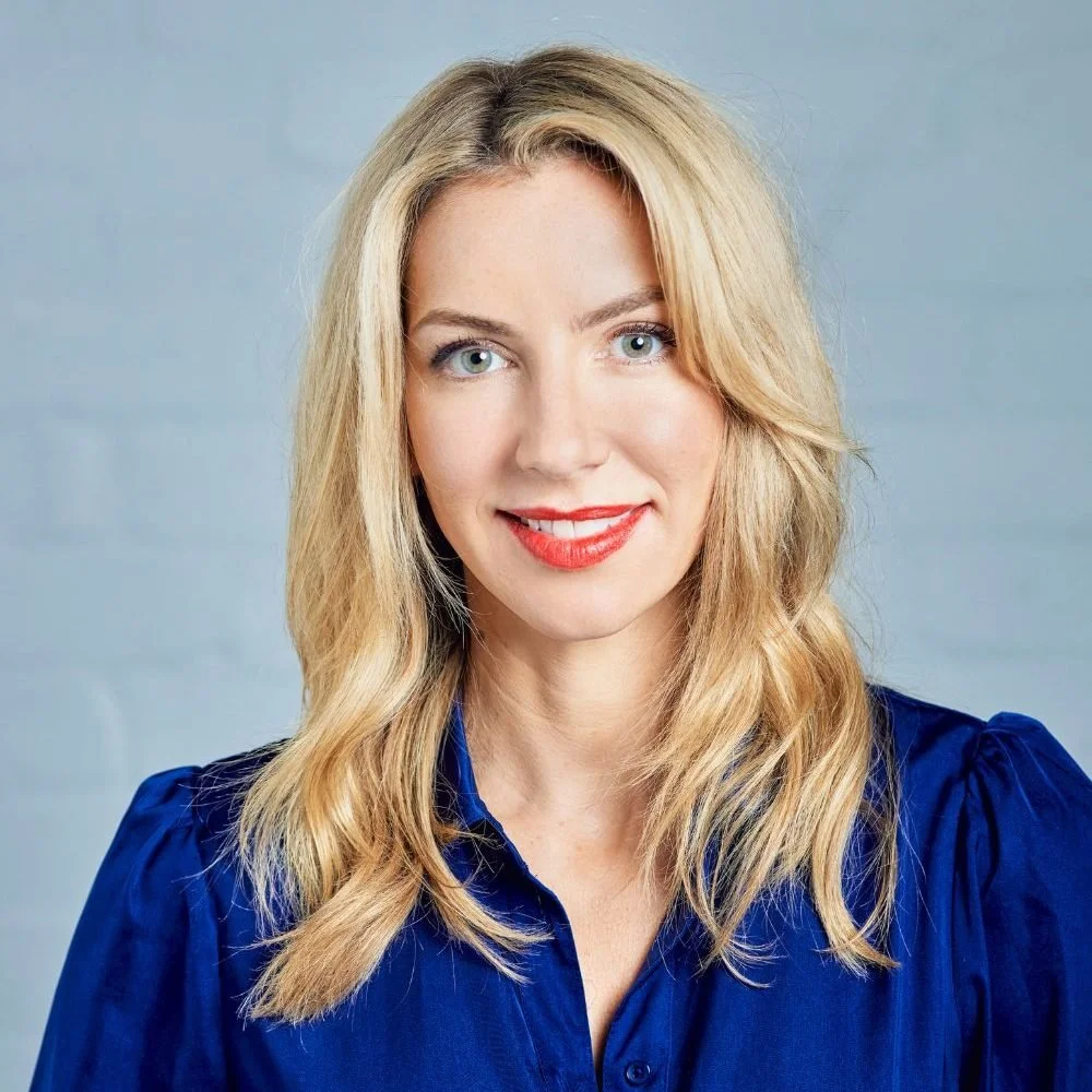 Liz Welsh. Portrait of a woman with blonde hair and blue eyes, wearing a blue blouse, smiling against a light gray background.