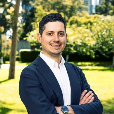 Nick Strongman from Sensum. A man in a dark suit with a white shirt, standing outdoors in a park with green trees and grass, smiling with arms crossed.