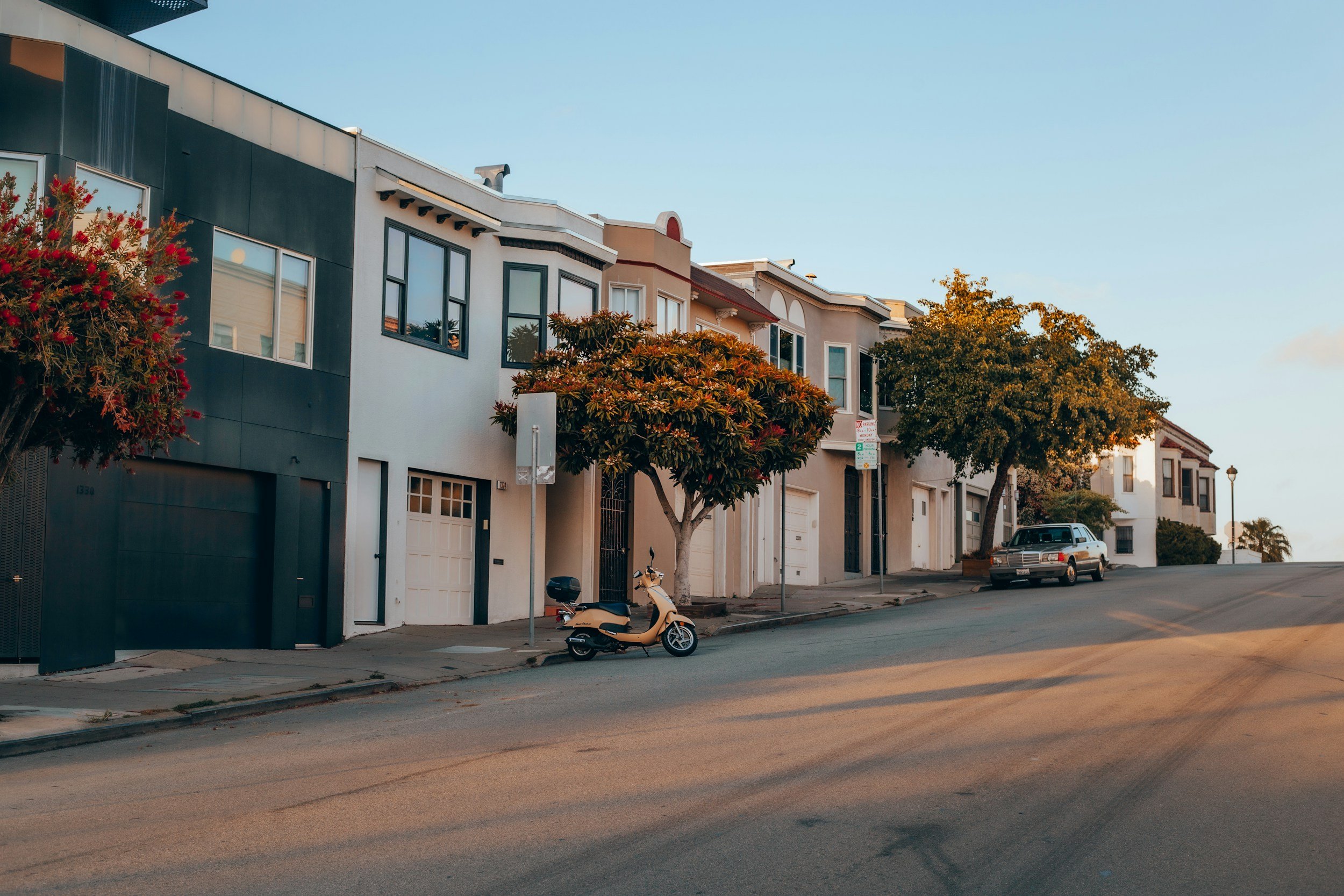 A quiet residential street with attached townhouses, trees, a parked car, and a scooter, under a clear blue sky during sunset.