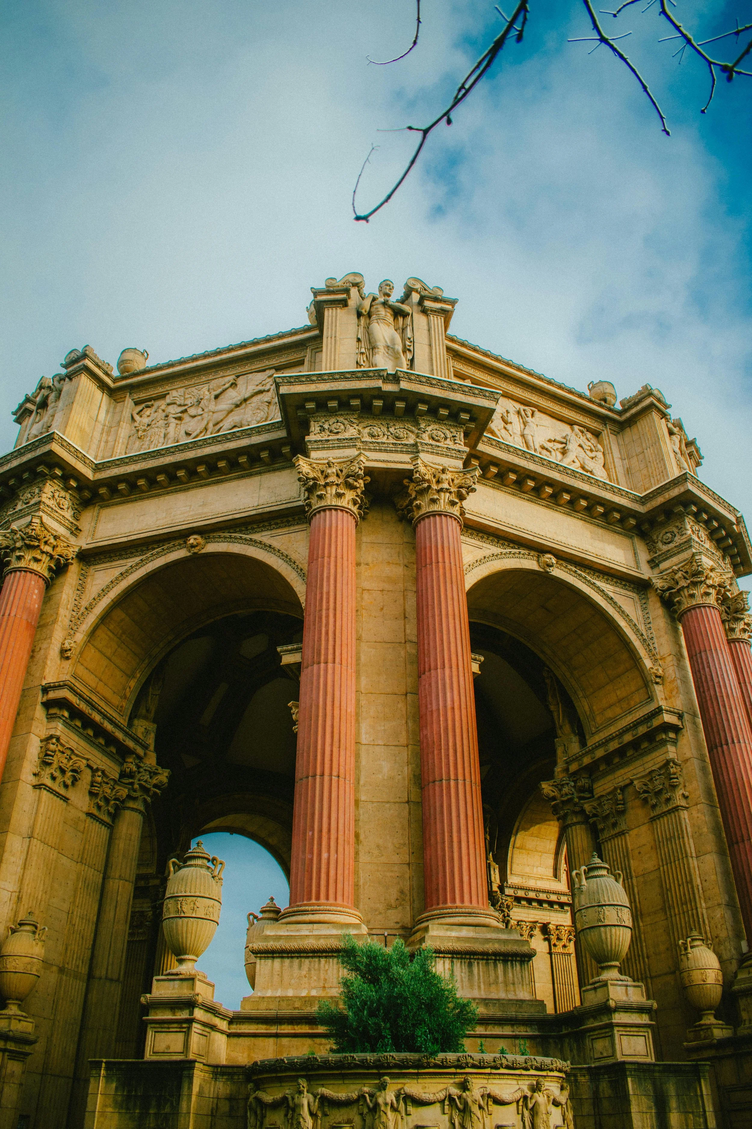 The image shows the top part of an architecturally elaborate building with Corinthian columns and detailed sculptures on the facade. The structure features a frieze with carved figures and a domed roof, with a statue of a woman at the center. The sky above is partly cloudy, and there are some tree branches visible at the top and green foliage at the bottom.