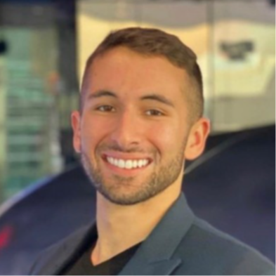 Portrait of a young man with short brown hair, smiling, wearing a dark blazer, in an indoor setting with blurred background.