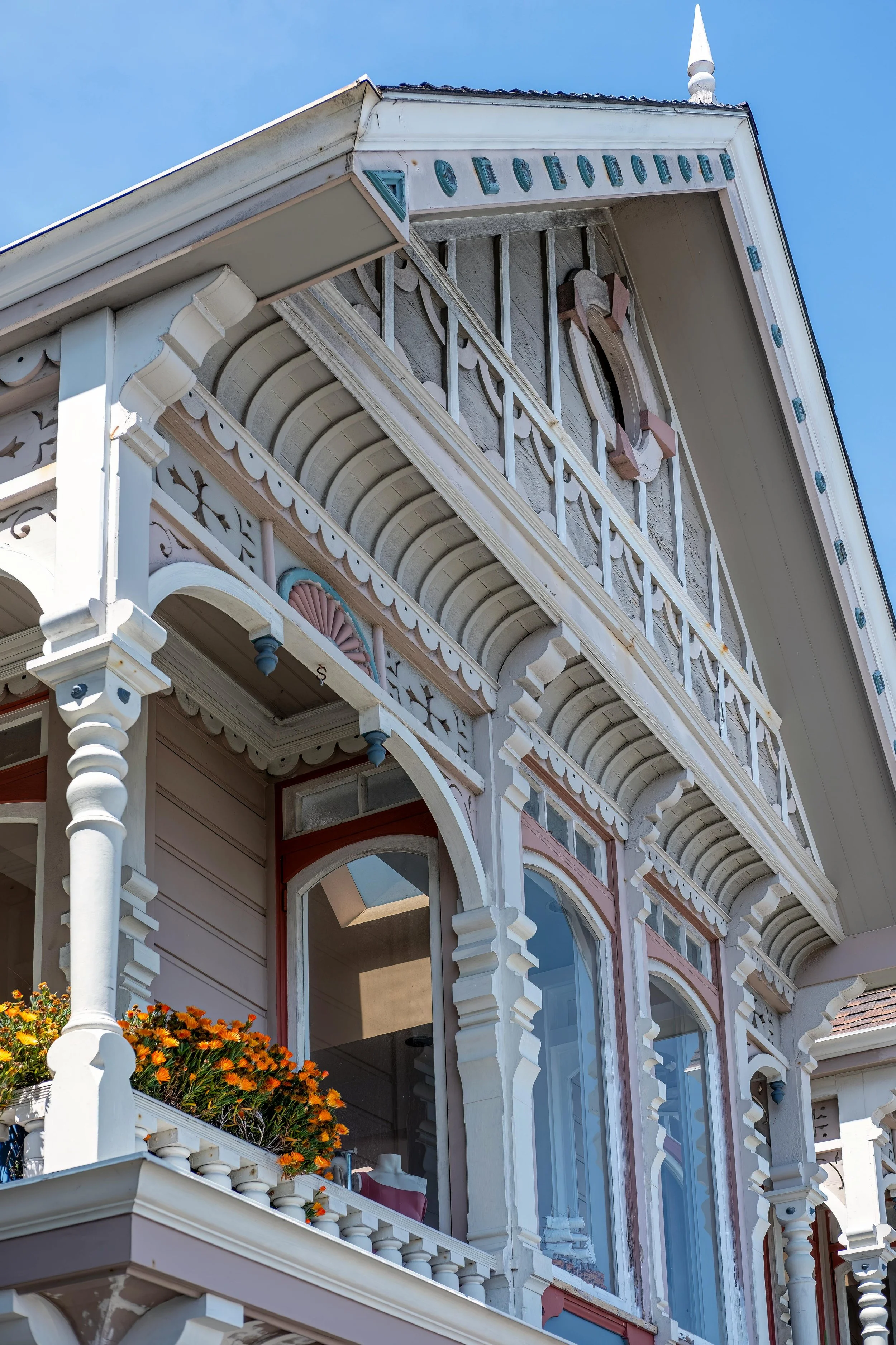 Close-up view of a Victorian-style house with intricate woodwork, decorative trim, and a porch railing, featuring a small balcony with orange flowers in a flower box, set against a clear blue sky.