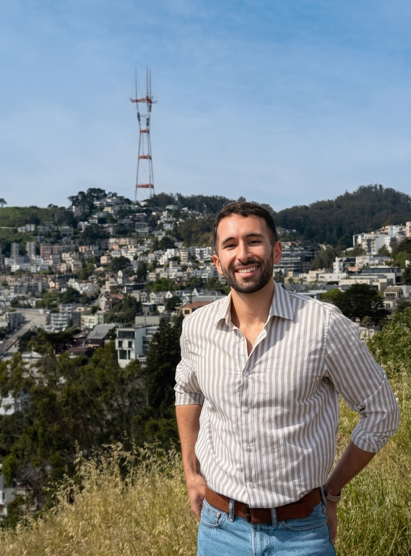 A smiling man in a striped shirt and jeans standing outdoors with a cityscape and a communication tower on a hill behind him.
