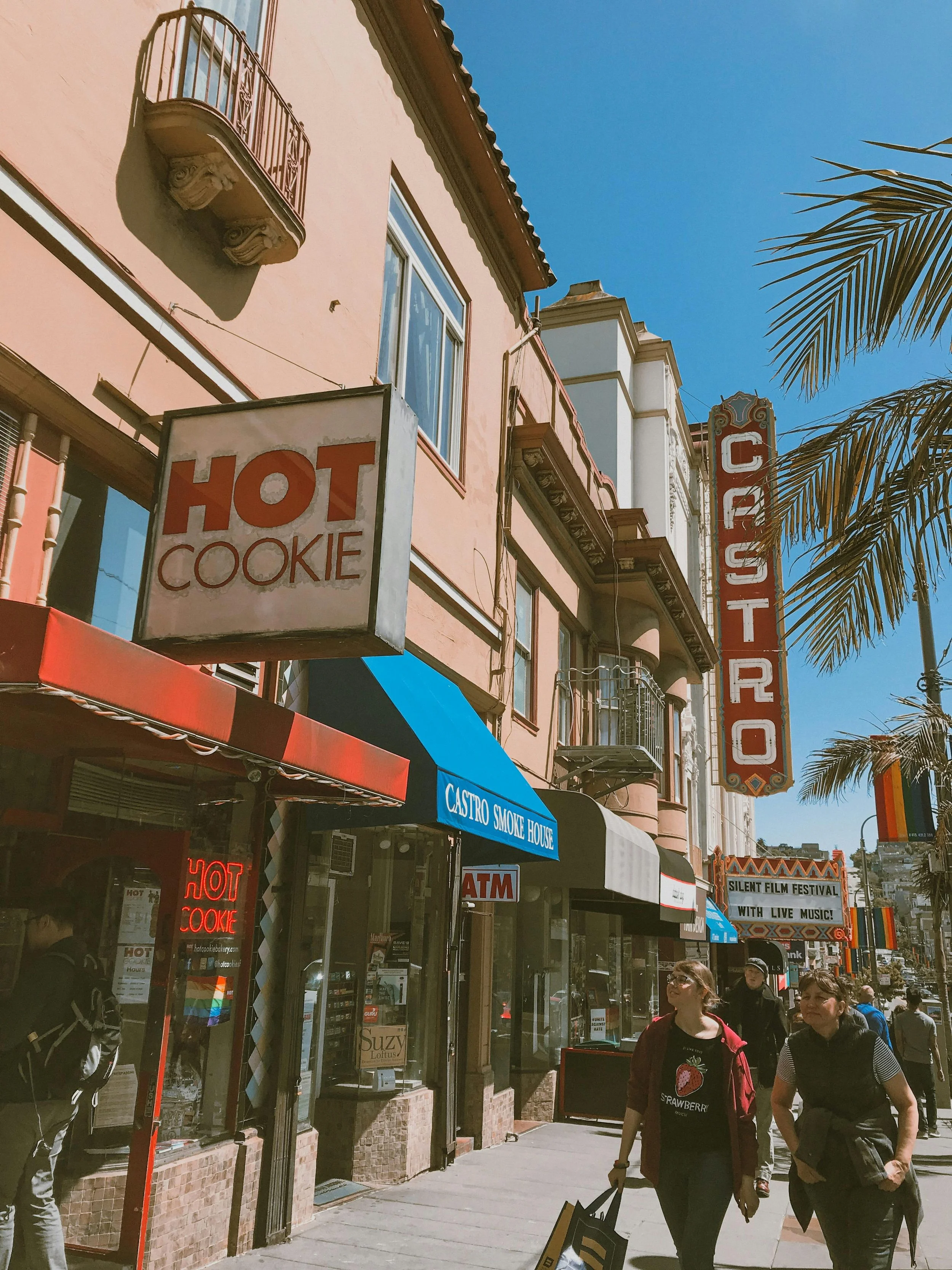 Pedestrians walking on a busy sidewalk in front of colorful buildings, including a bakery with signs for hot cookies and a smokehouse, under a clear blue sky with a palm tree on the right.