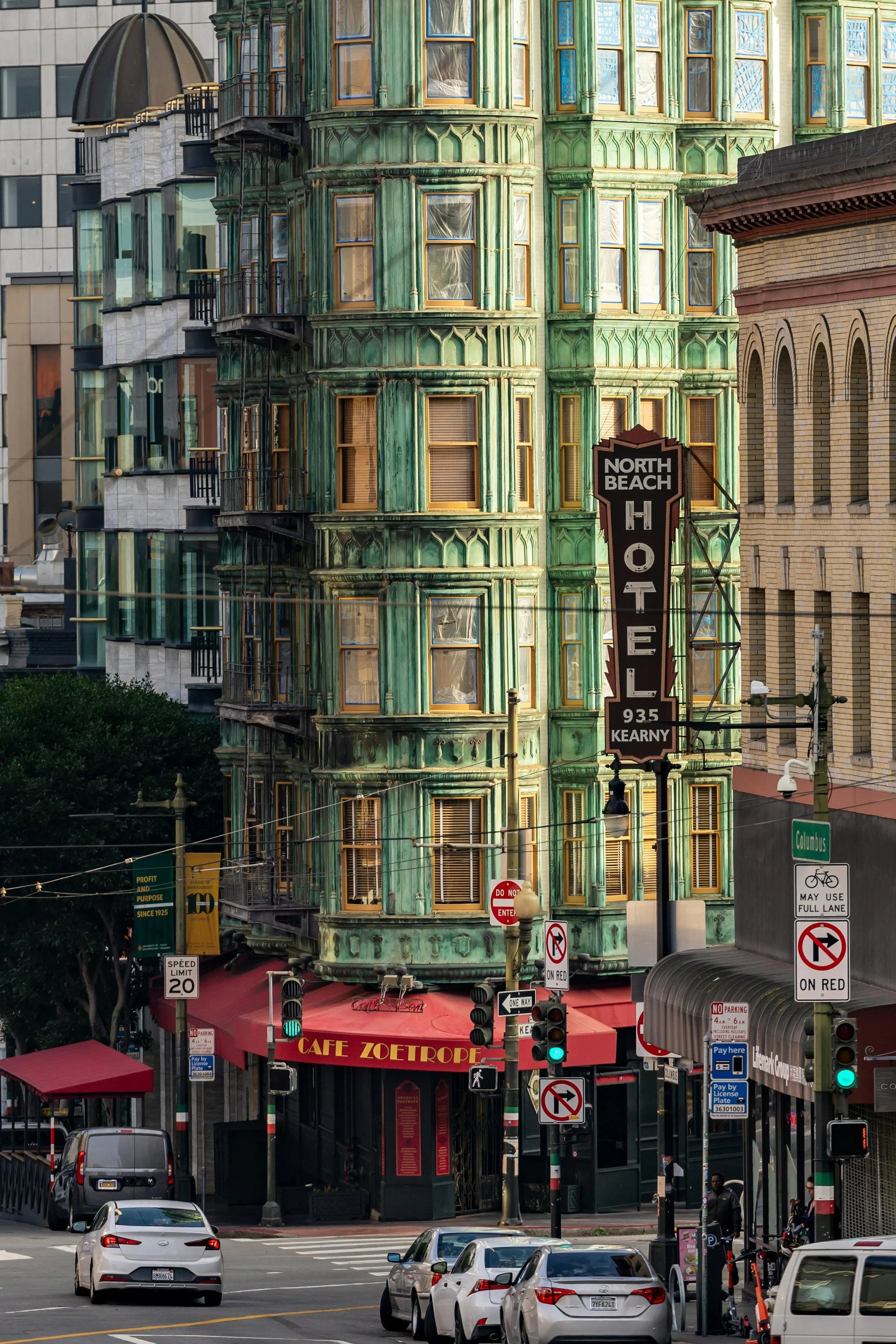 Street scene in an urban area featuring a historic green building with ornate architecture, a storefront sign for North Beach Hotel, and cafes with outdoor seating. Cars and pedestrians are visible, along with street signs and traffic lights.