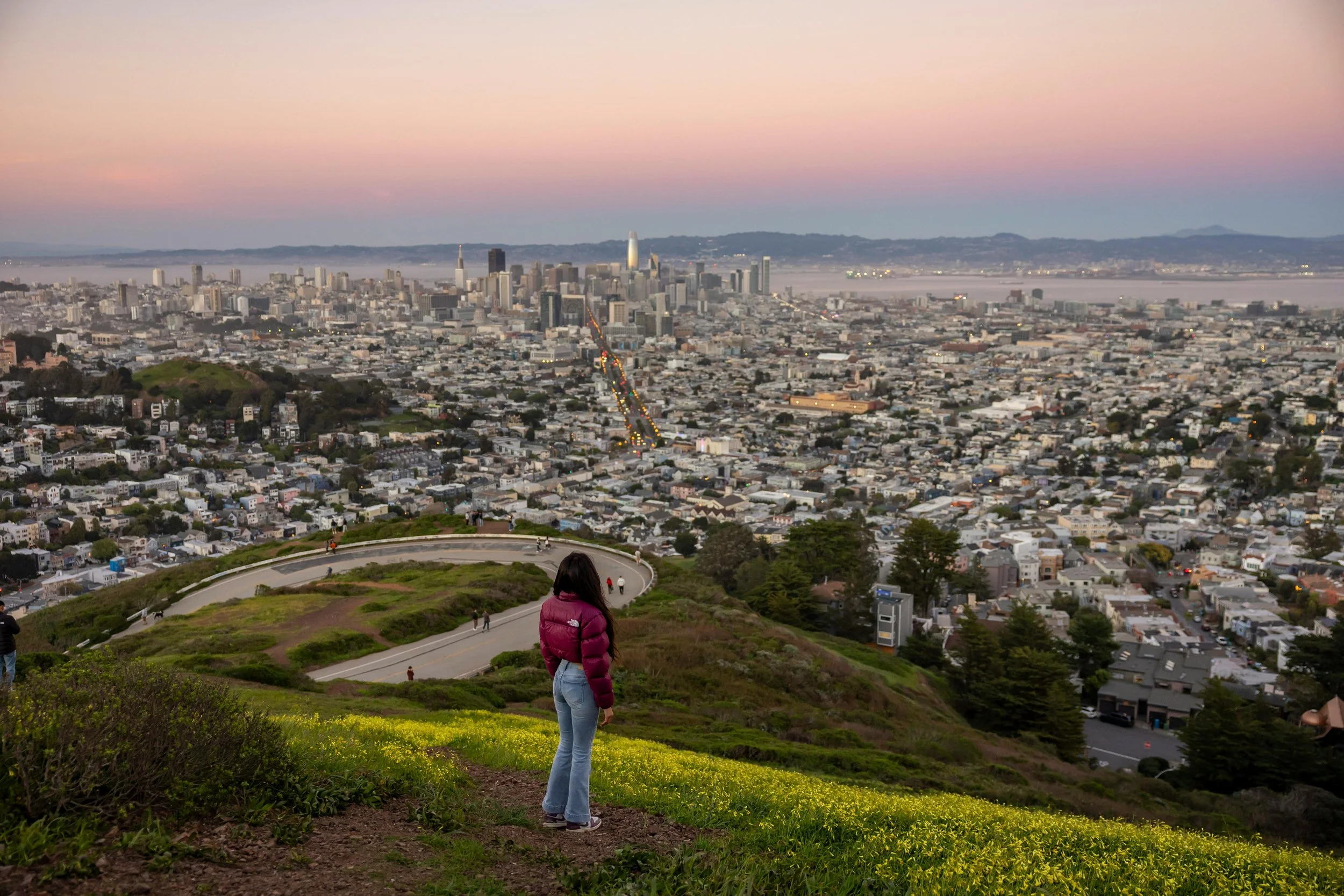 A woman in a purple jacket and jeans standing on a grassy hill overlooking San Francisco skyline at dusk.