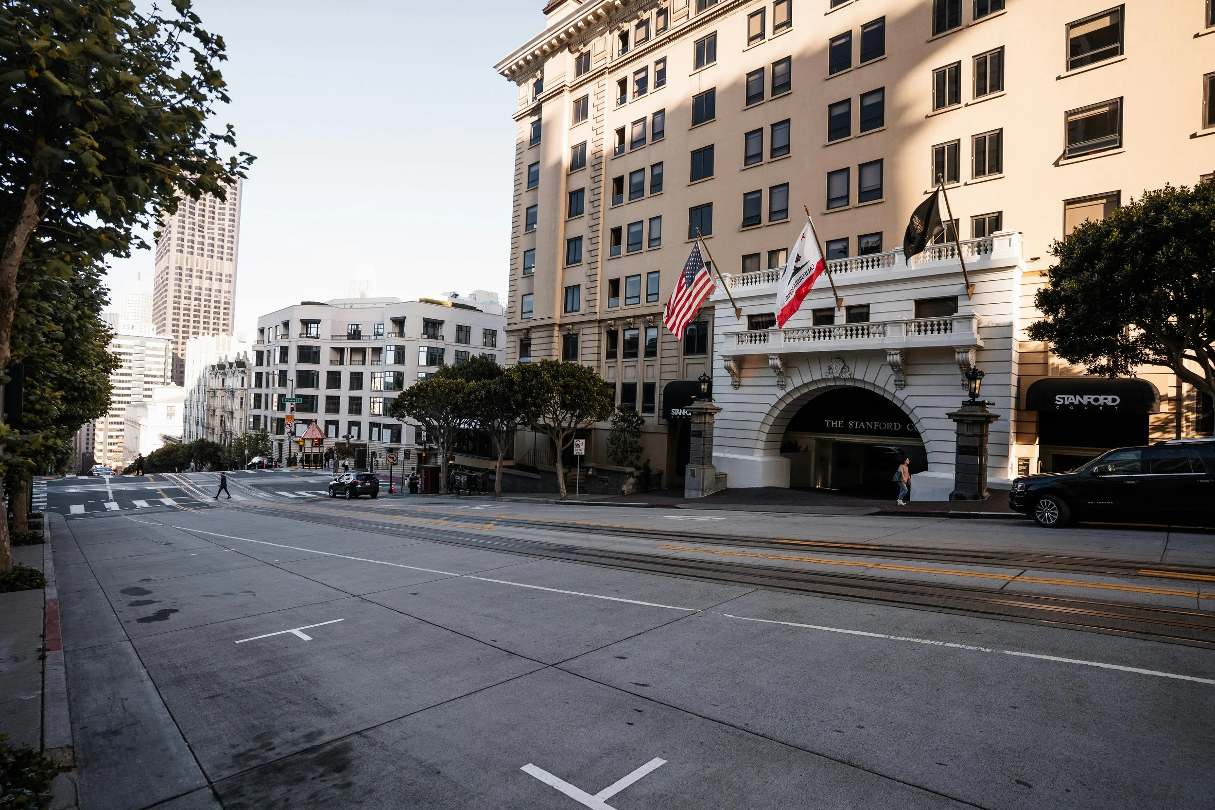 A city street scene with a large white multi-story building with flags, trees, and cars parked along the street, and a few pedestrians walking.