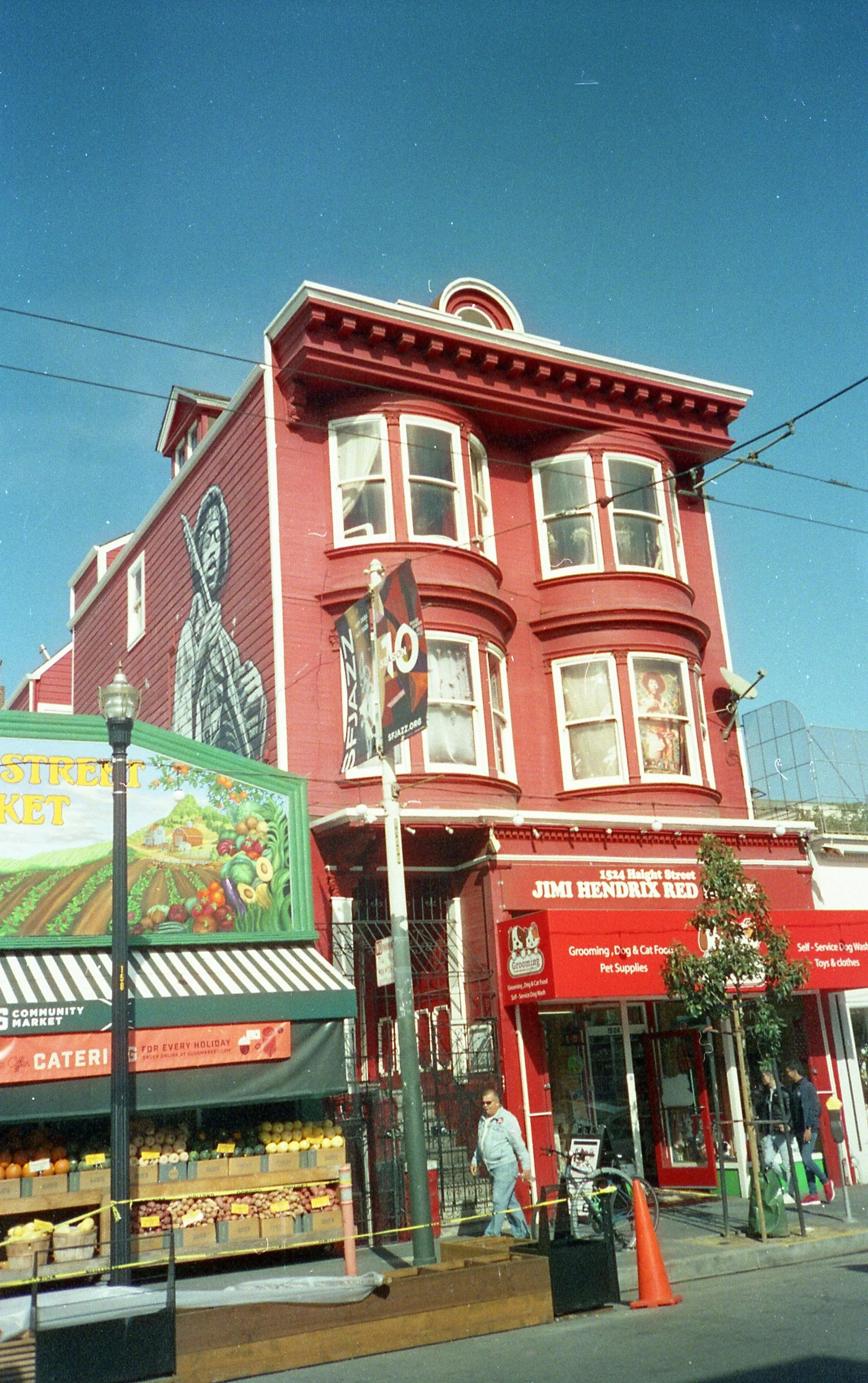 A red multi-story building with an animated figure mural on its side, street market stalls in front, and people walking along the sidewalk.