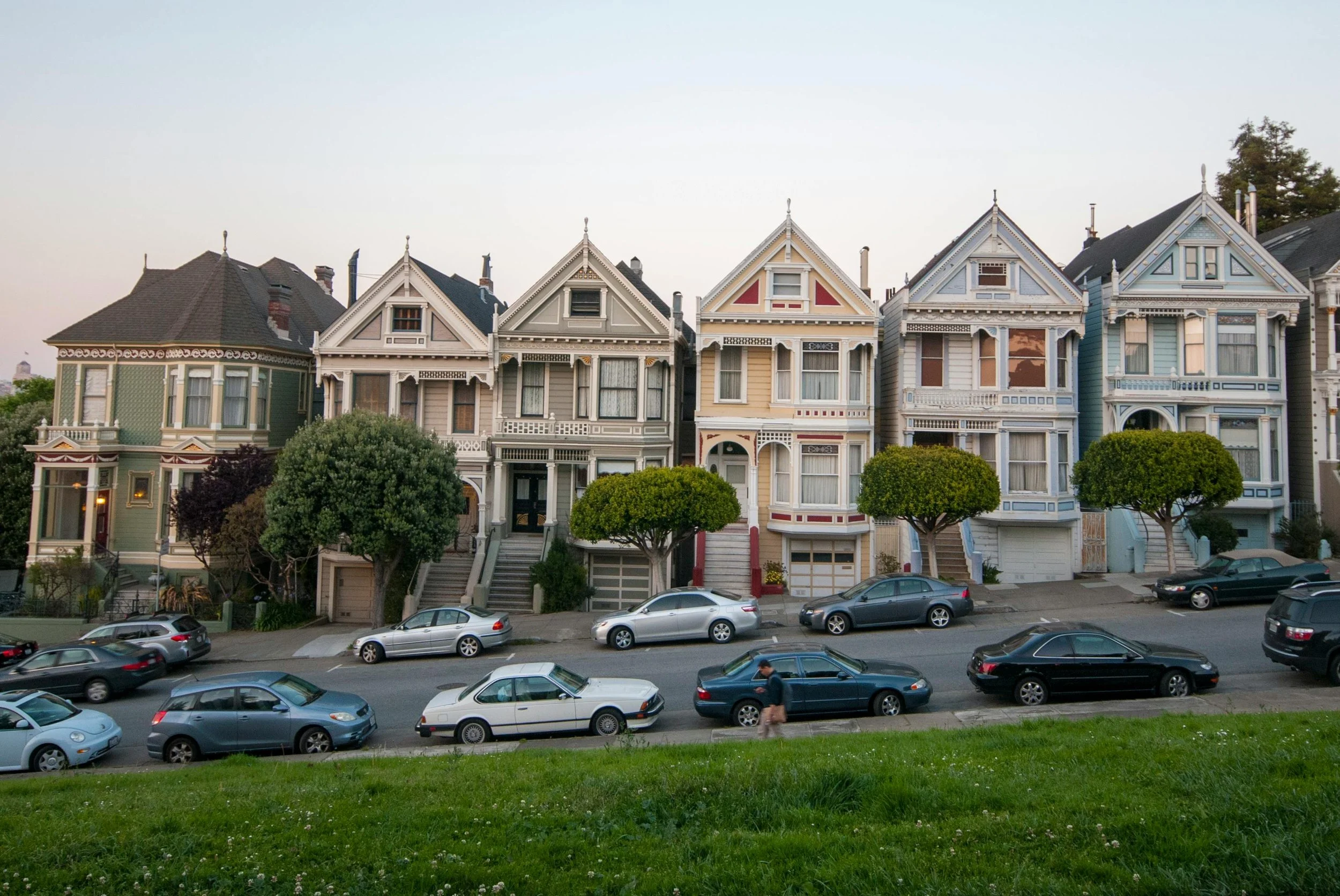 A row of colorful Victorian-style houses with front porches, set behind neatly trimmed trees, on a street with various parked cars and a person walking.