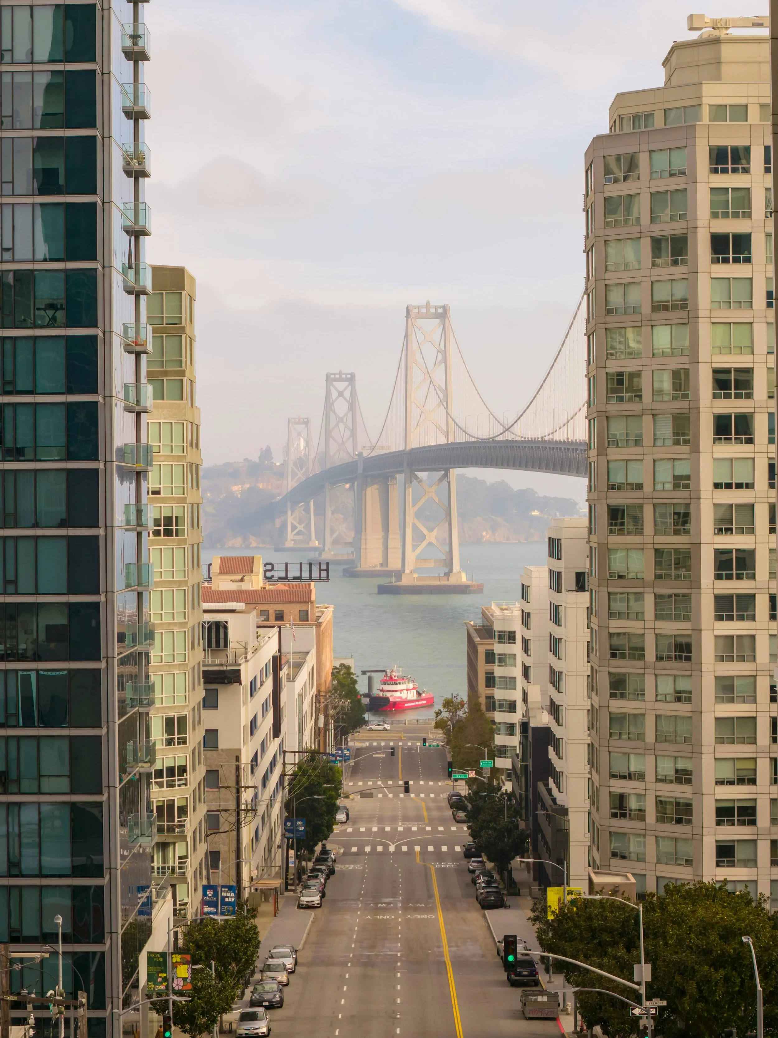 View of a city street flanked by high-rise apartment buildings, leading to the San Francisco-Oakland Bay Bridge in the distance, with a ferry boat on the water.