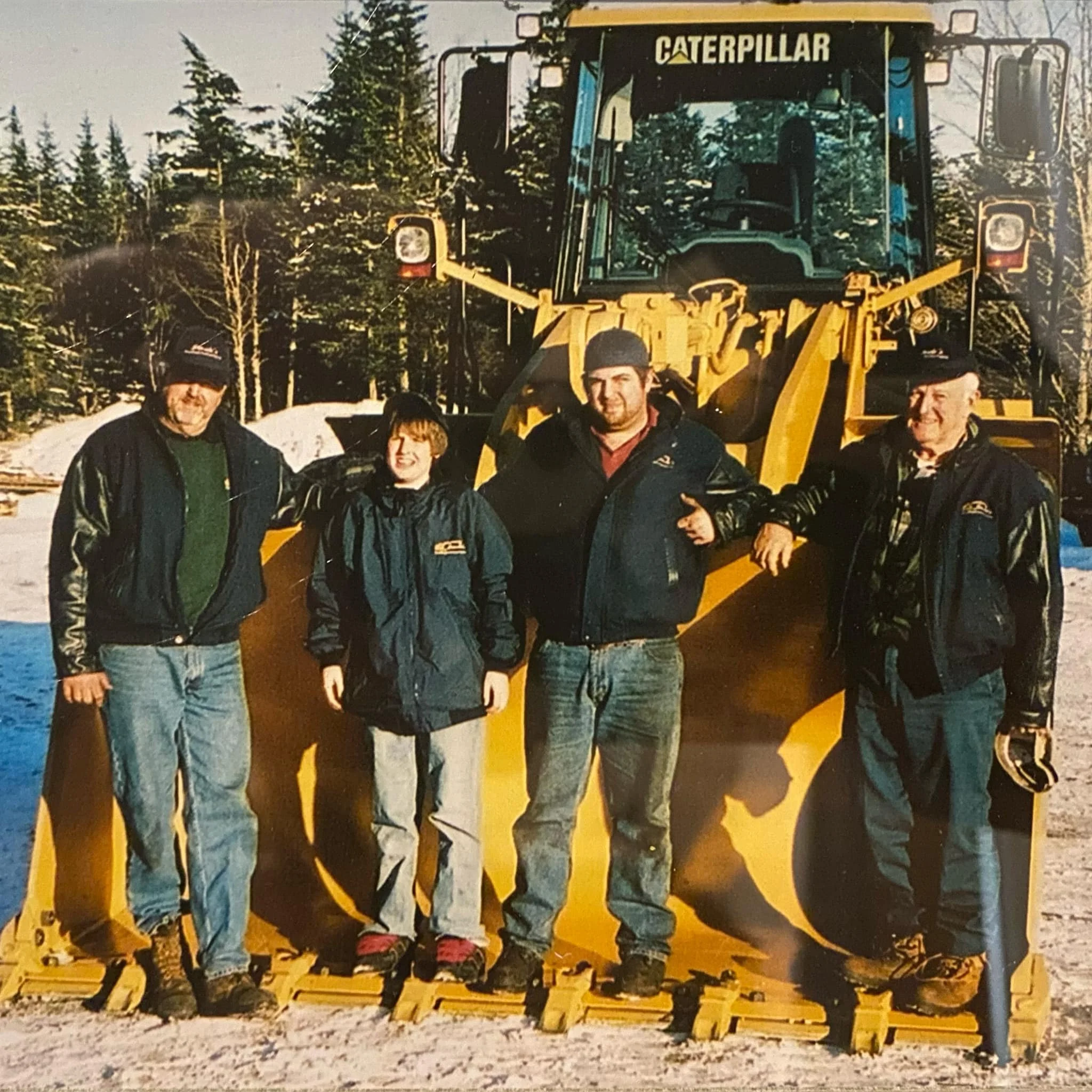 Group of people standing on snow, posing in front of a yellow Caterpillar snow removal vehicle, with background of trees and snowfall.