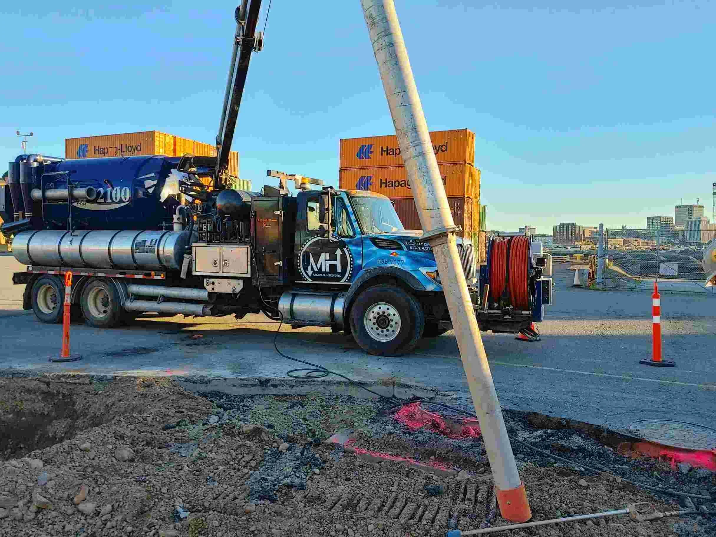 Construction site with a truck and crane lifting a large pipe, orange cones, and city buildings in the background.