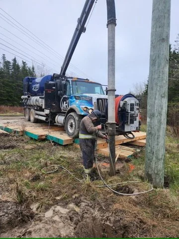 Worker operating drilling equipment on roadside near utility pole, with a large blue truck nearby.