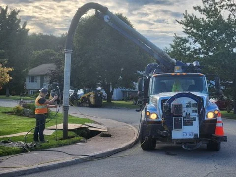 A worker in orange safety vest and helmet operating a vacuum truck to clean a storm drain on a residential street.