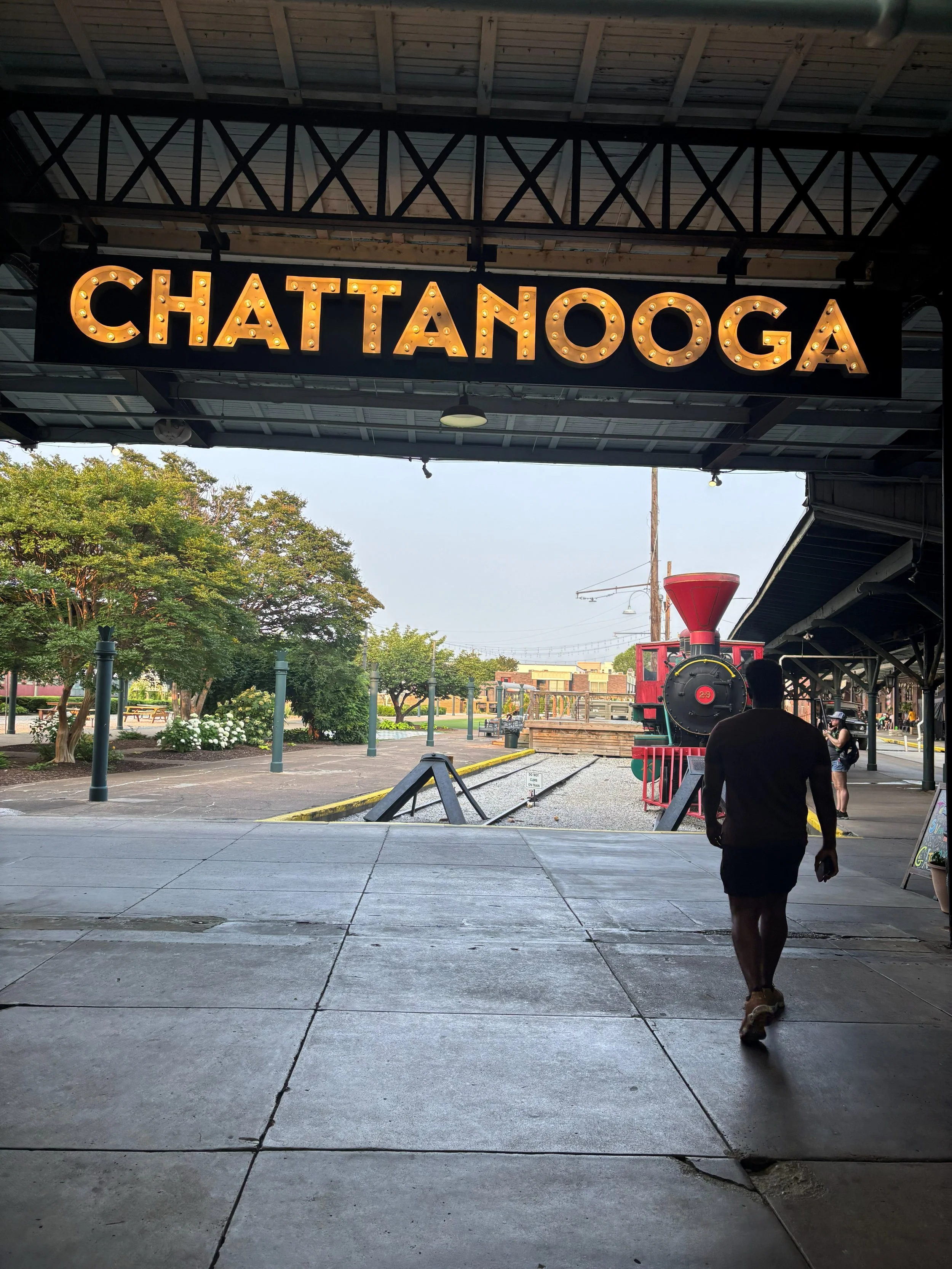 View of a train station platform with a large illuminated sign spelling 'CHATTANOOGA', and a vintage red locomotive in the background. A person is walking away from the camera, and there are trees and a city street scene beyond the platform.