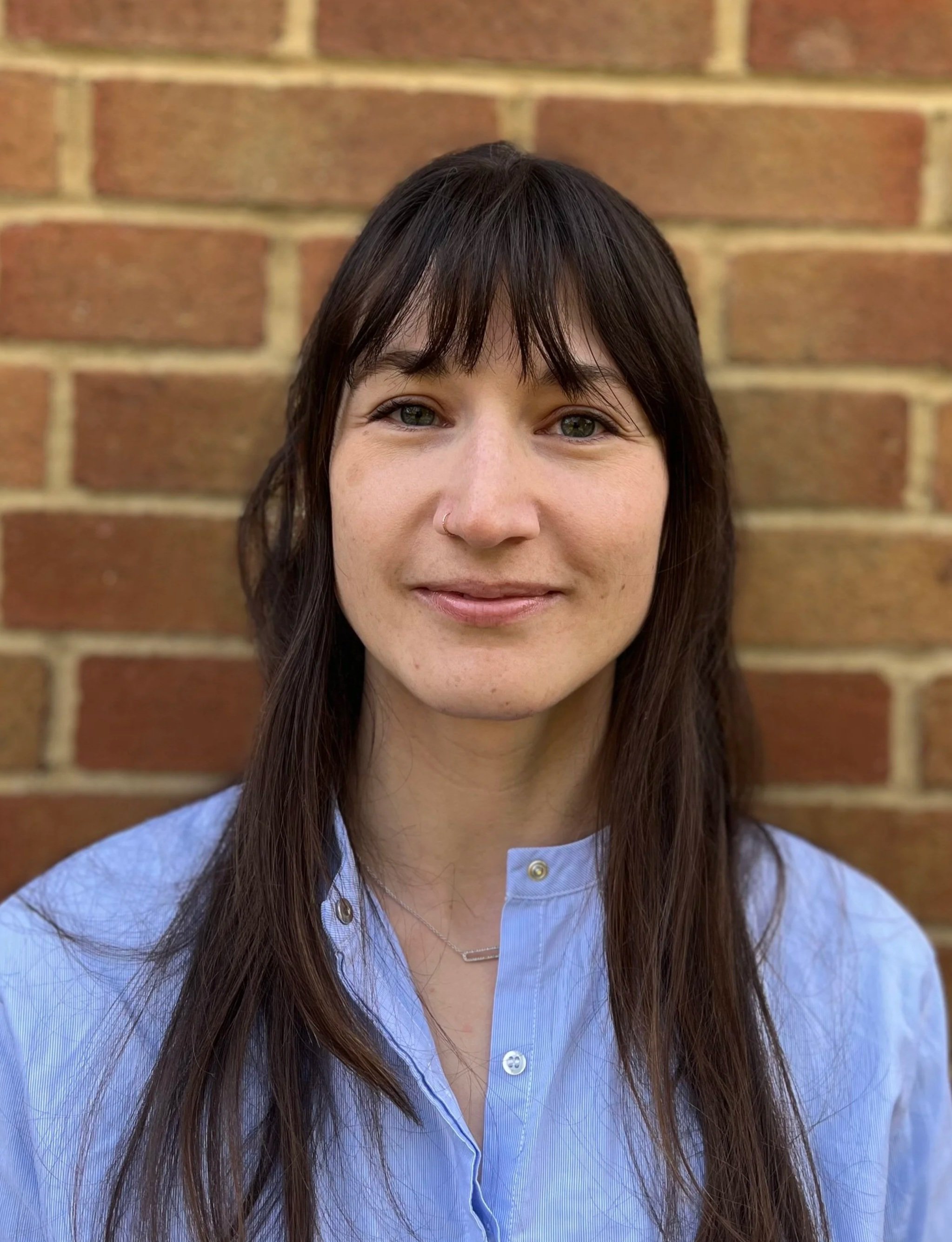 A woman with long brown hair and a nose ring smiling in front of a brick wall, wearing a light blue shirt.