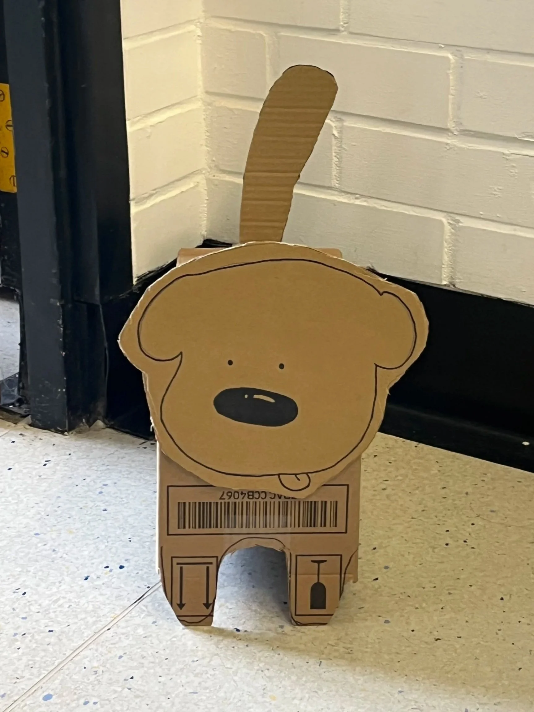Cardboard box decorated as a dog with a brown dog face and ears, placed on a tiled floor in front of a white brick wall.