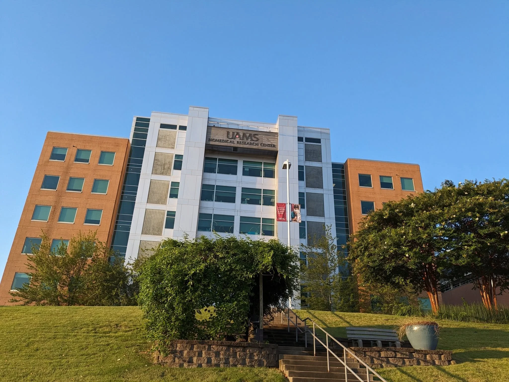A modern multi-story building with a mix of white and brown exterior, featuring numerous windows. The building is labeled 'UAMS Biomedical Research Center.' In the foreground, there is a lush green lawn with a small bush, a staircase with metal railings, and a large white planter at the bottom right.
