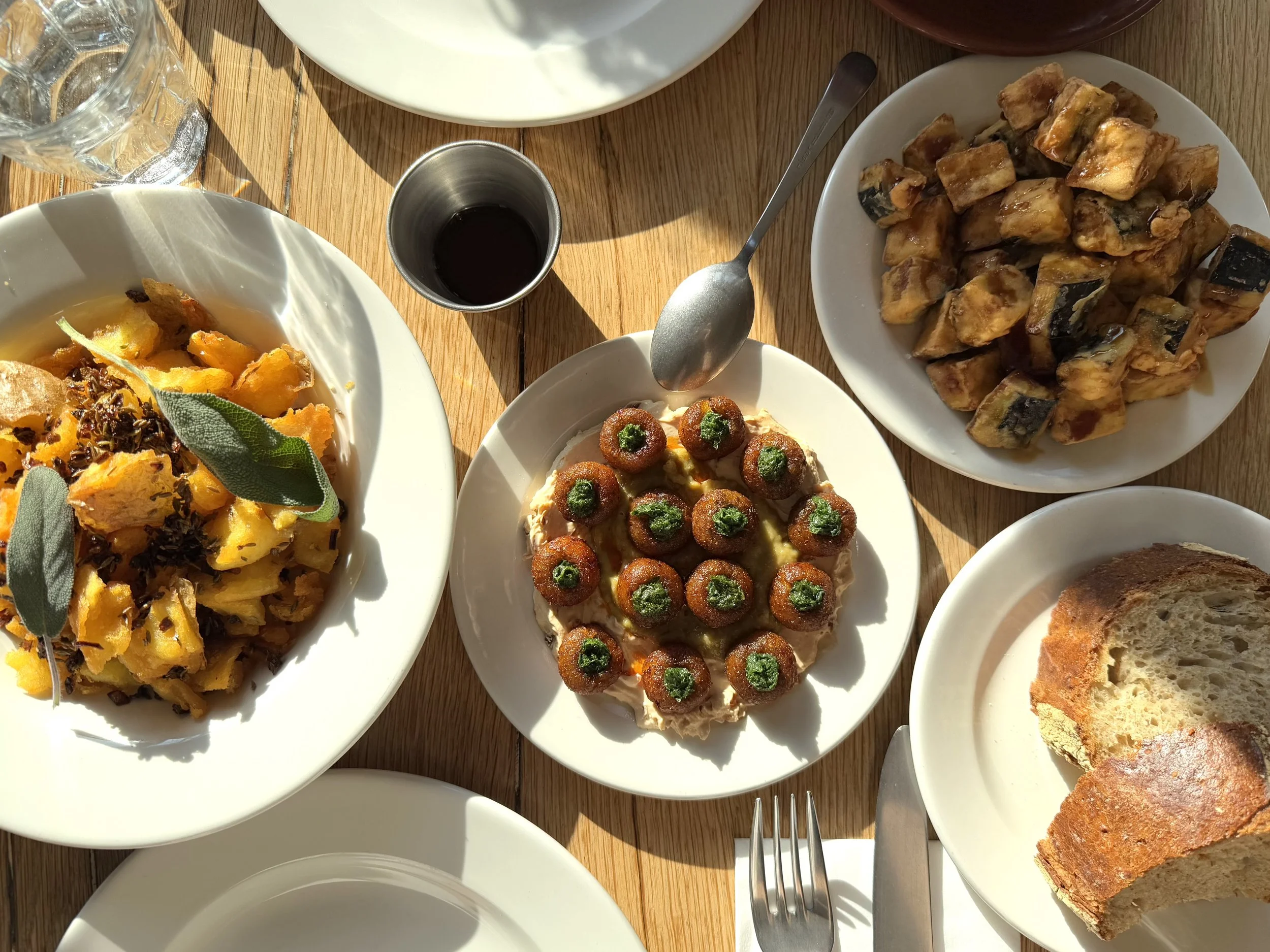 Assorted dishes on a wooden table, including a bowl of roasted squash with sage leaves, crispy croquettes with green herbs, a plate of sautéed eggplant, slices of bread, and a small glass of red wine.