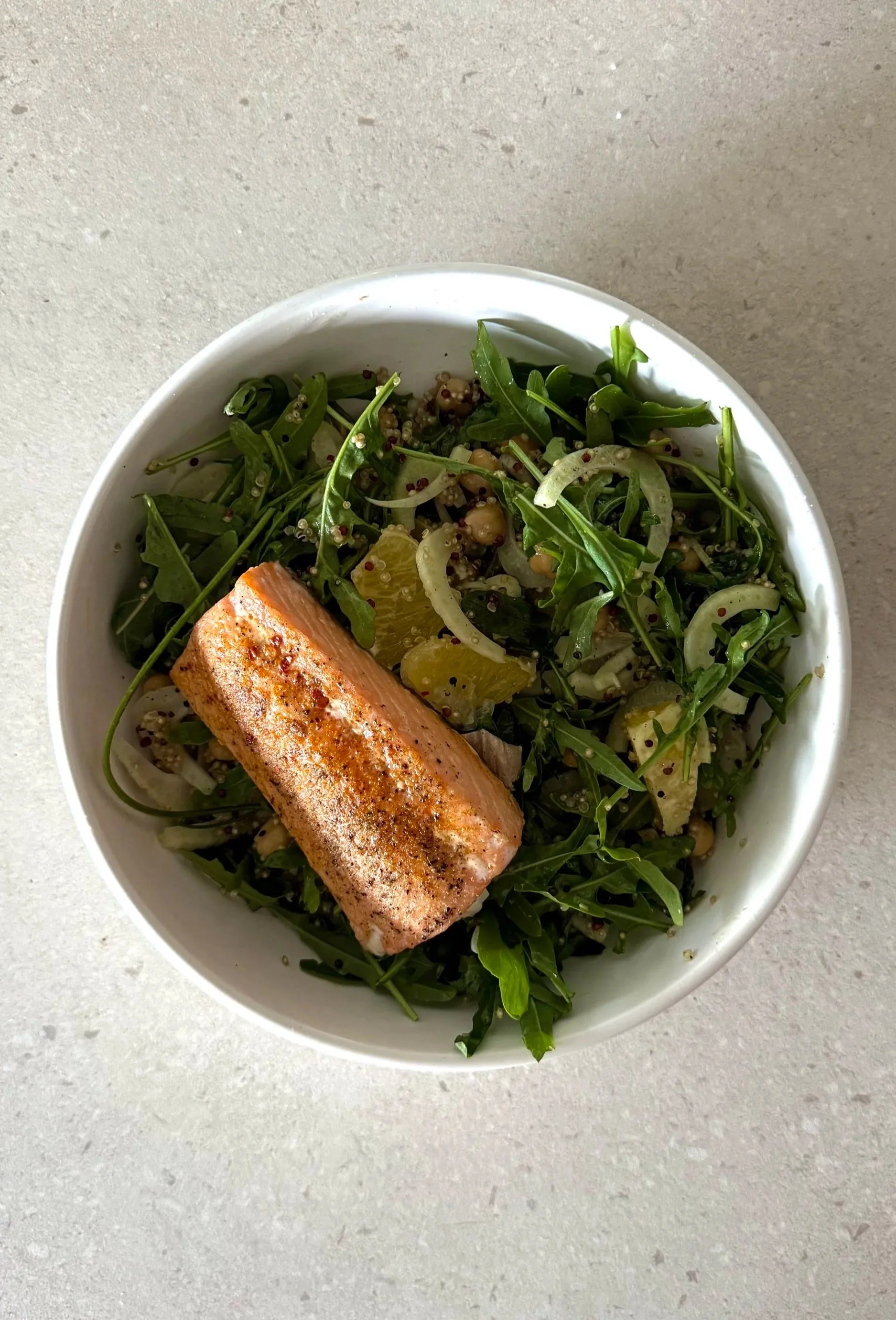 A bowl of salad with arugula, lemon slices, and a seasoned salmon fillet on top.