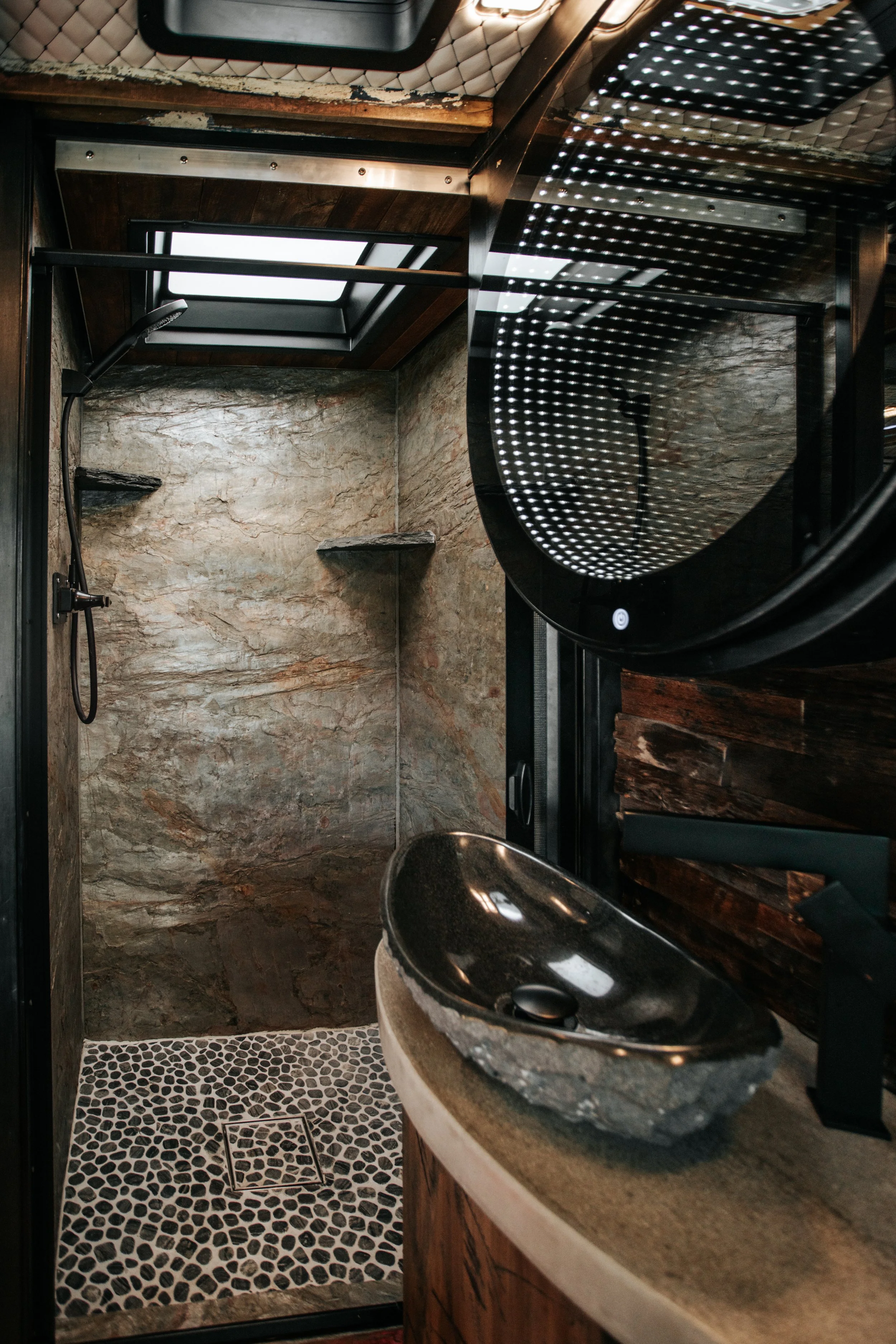 Small bathroom with a pebble stone floor, stone shower walls, a rainfall showerhead, a skylight window, a dark wood vanity with a black vessel sink, and a large circular mirror reflecting the ceiling lights.