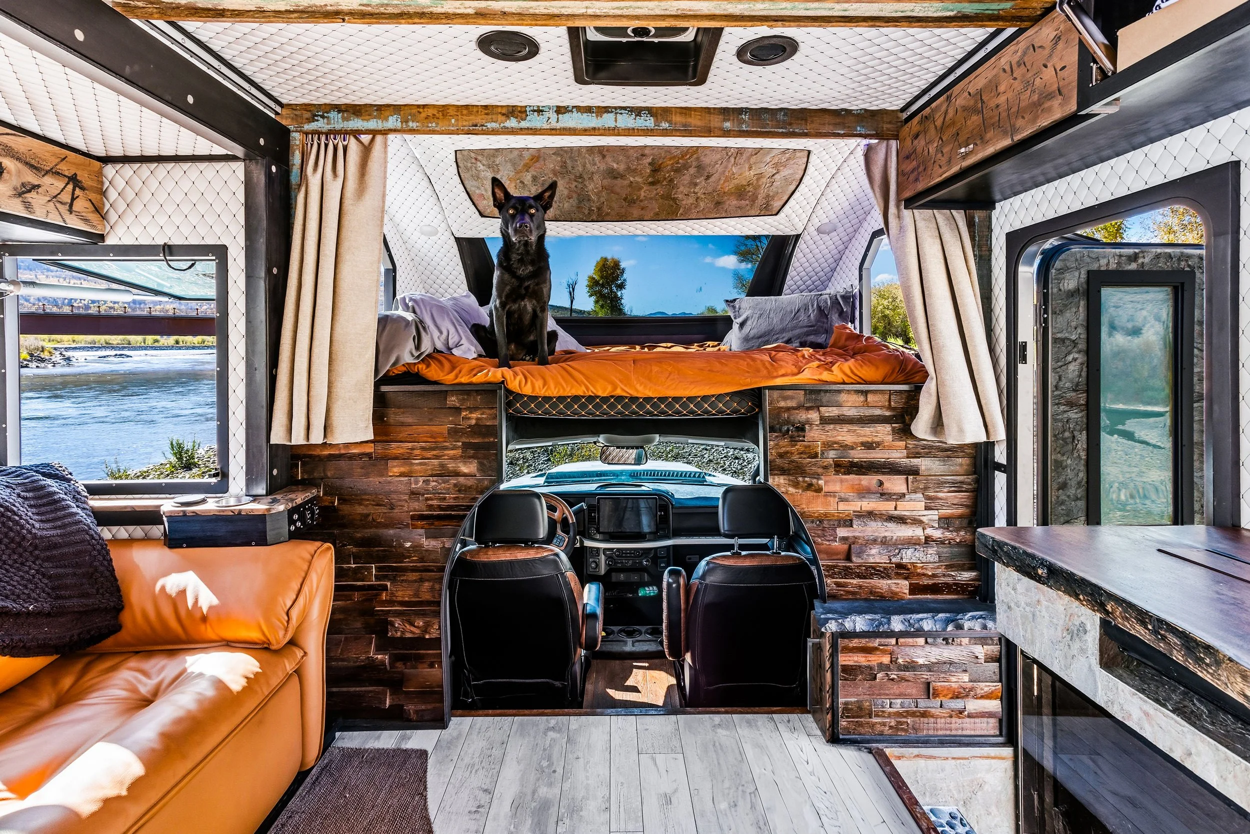 Interior of a compact living space inside a converted vehicle or camper with wood paneling, a bed with orange bedding, a black dog sitting on the bed, windows showing a river and trees in the background, a seating area with an orange sofa, and a kitchen counter with a marble top.