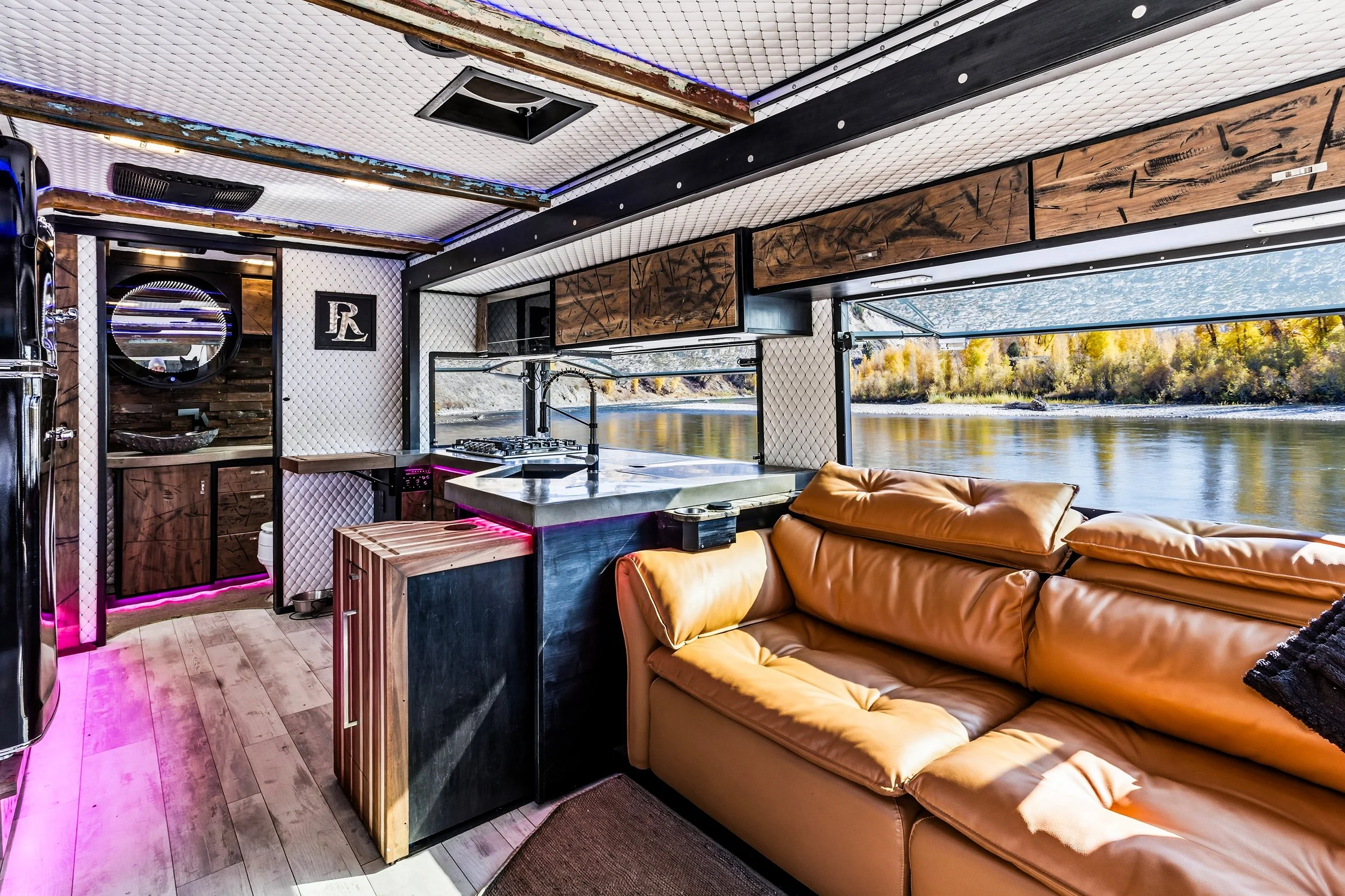 Interior of a houseboat with a kitchen and a tan leather sofa, overlooking a river and trees with autumn foliage, with large windows letting in natural light.