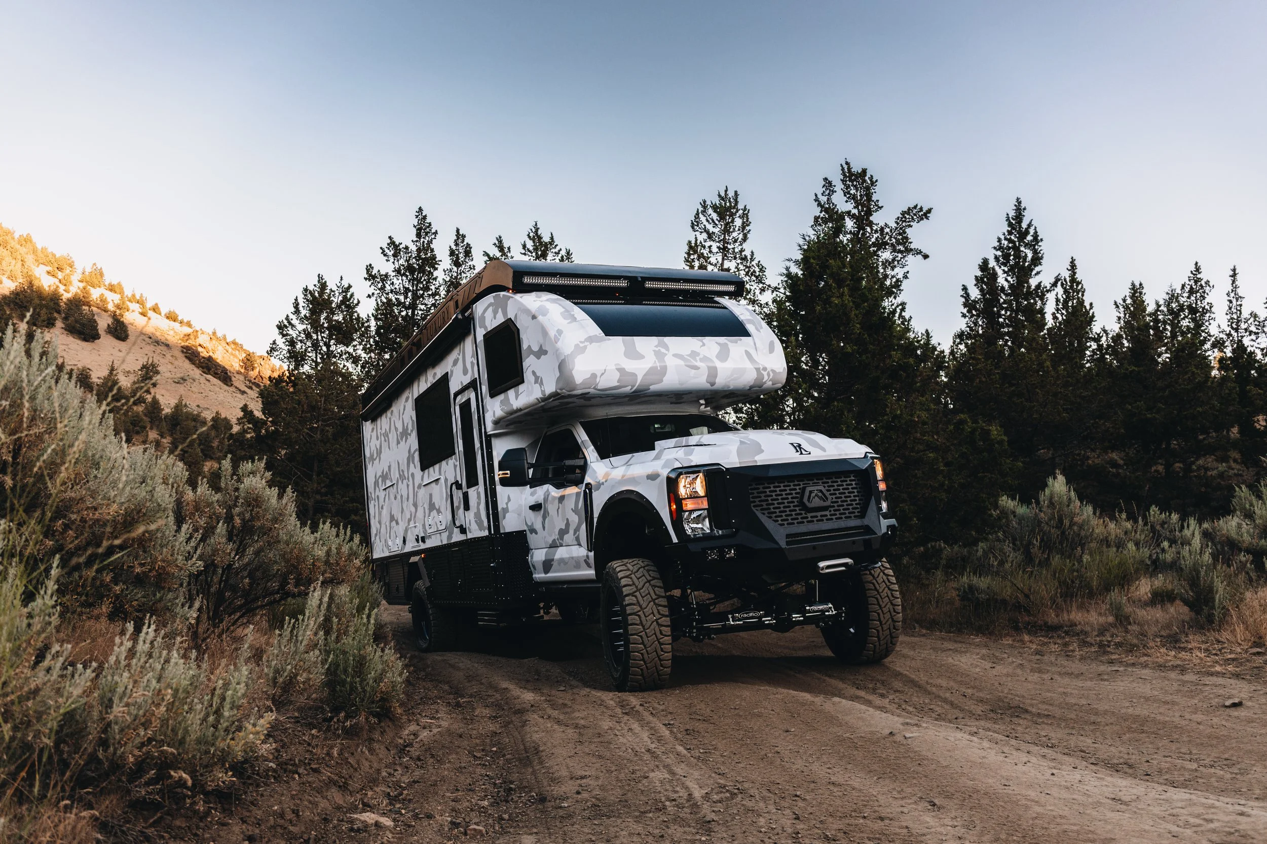 A white camper van with camouflage design parked on a dirt trail in a wooded area with tall pine trees and mountains in the background.