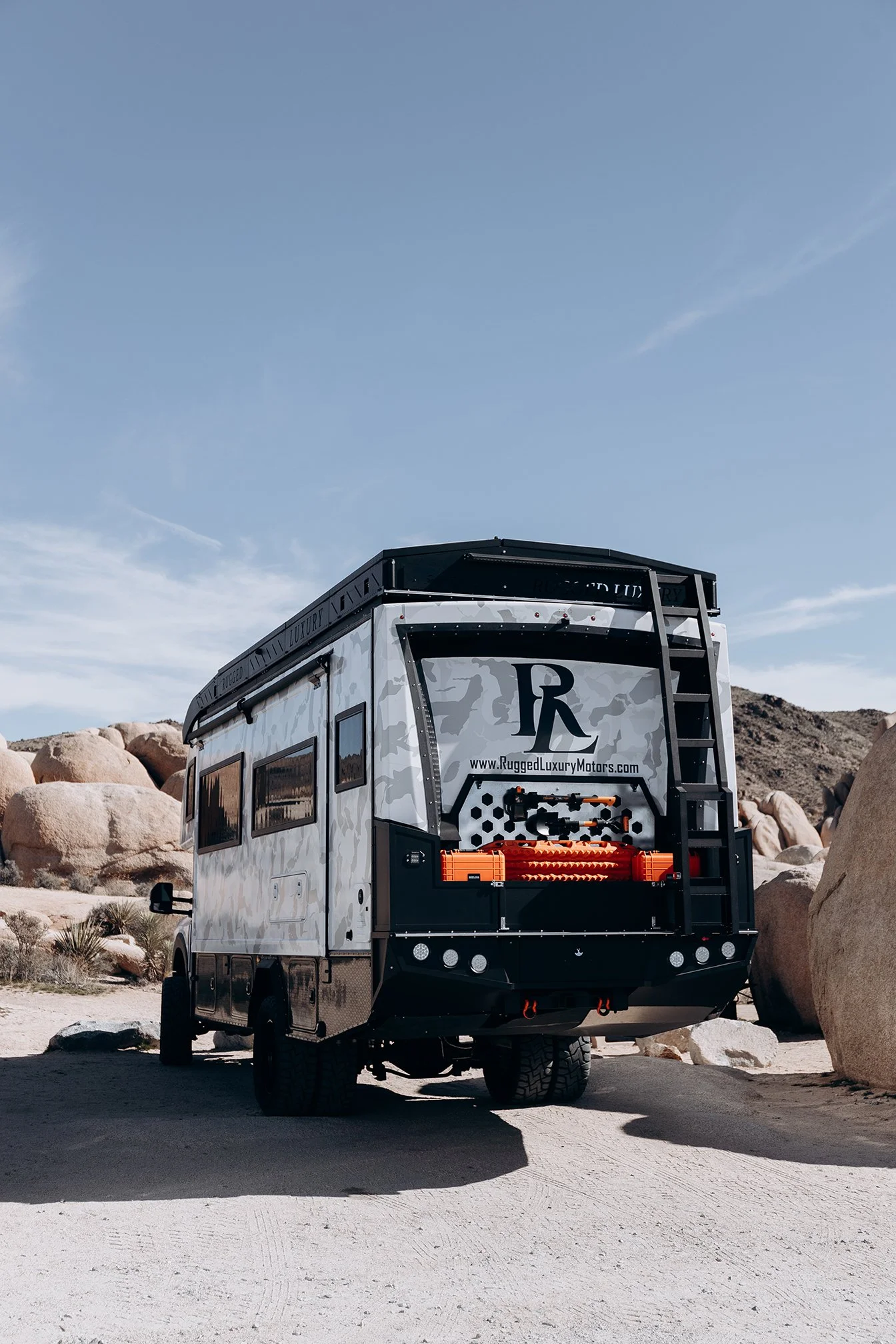 A luxury RV parked in a desert landscape with large rocks and dry vegetation, under a clear blue sky.