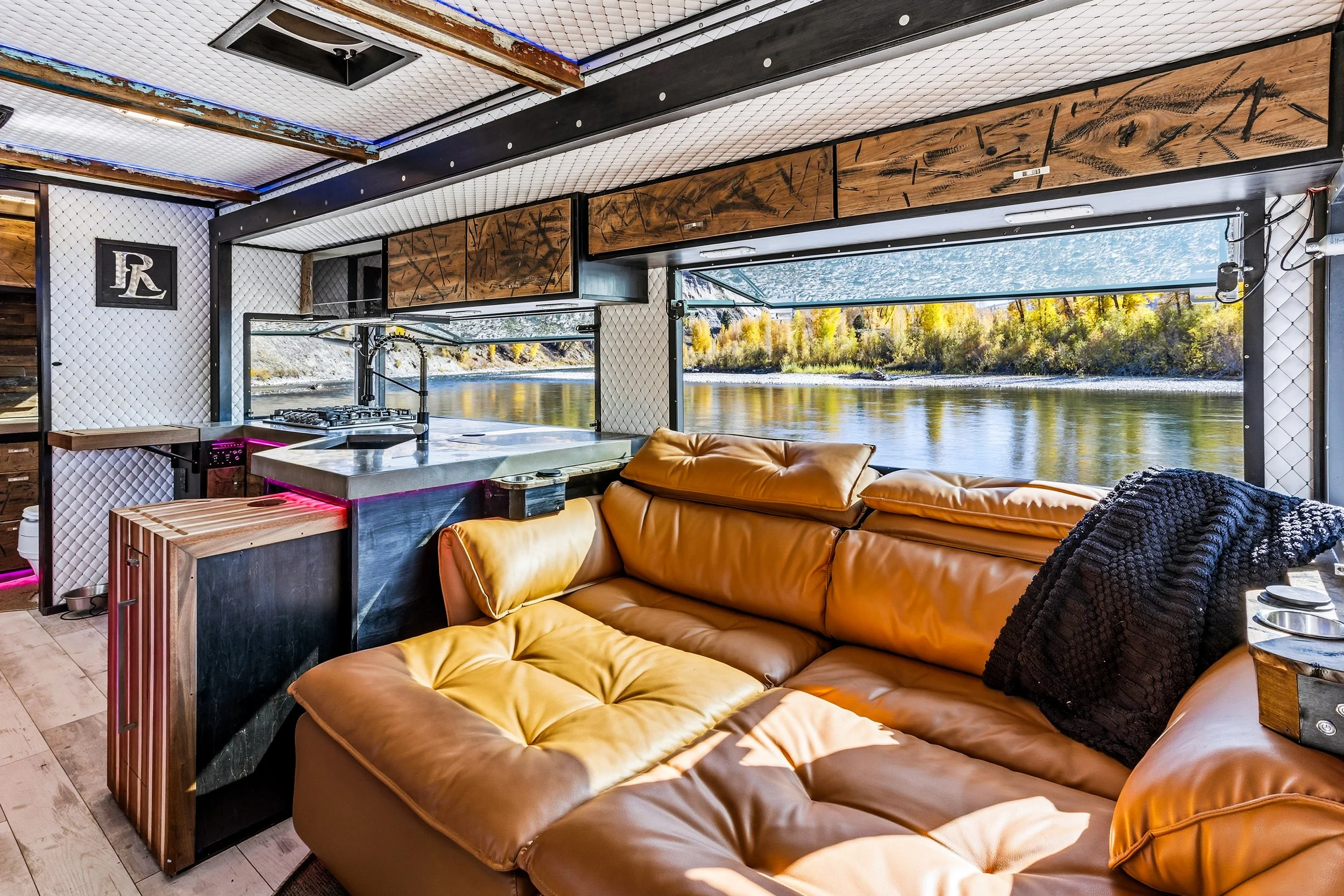 Interior of a houseboat with a brown leather sectional sofa, a black blanket, a small wooden side table, a kitchen area with a marble countertop, and large windows showing a river and autumn trees outside.